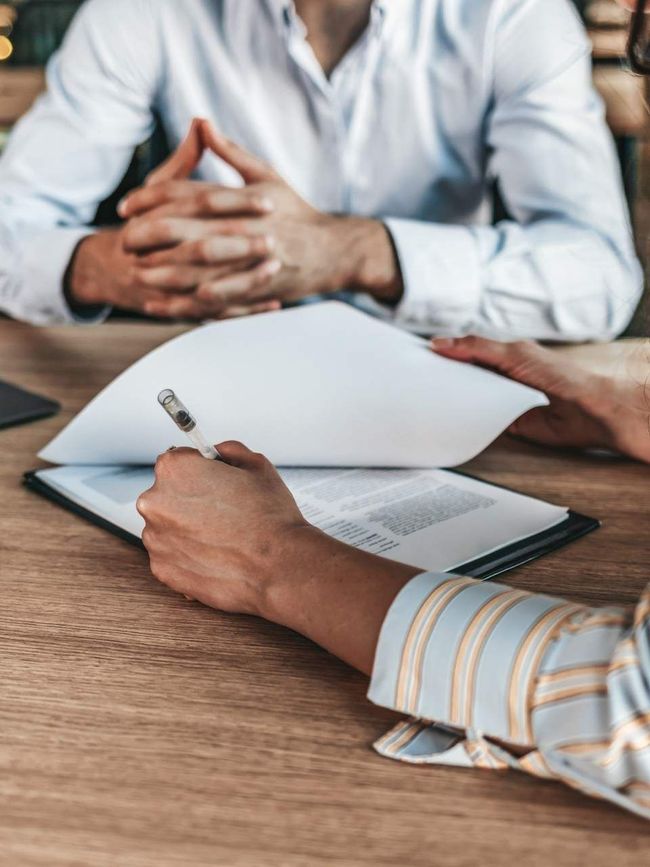 Hands holding a contract, reviewing documents at a wooden table with another person.