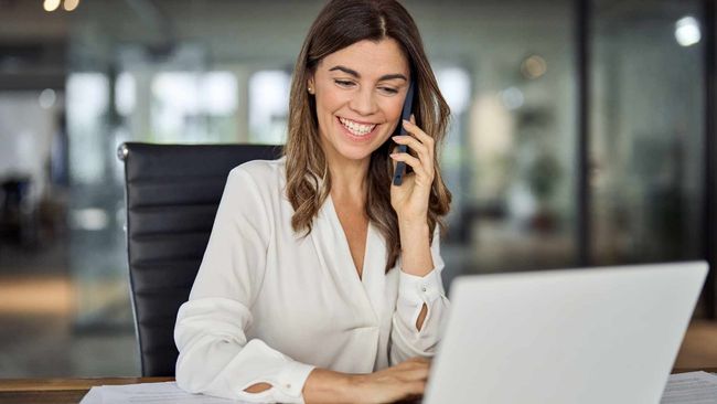 Woman smiles while on a phone call, using a laptop in an office setting.