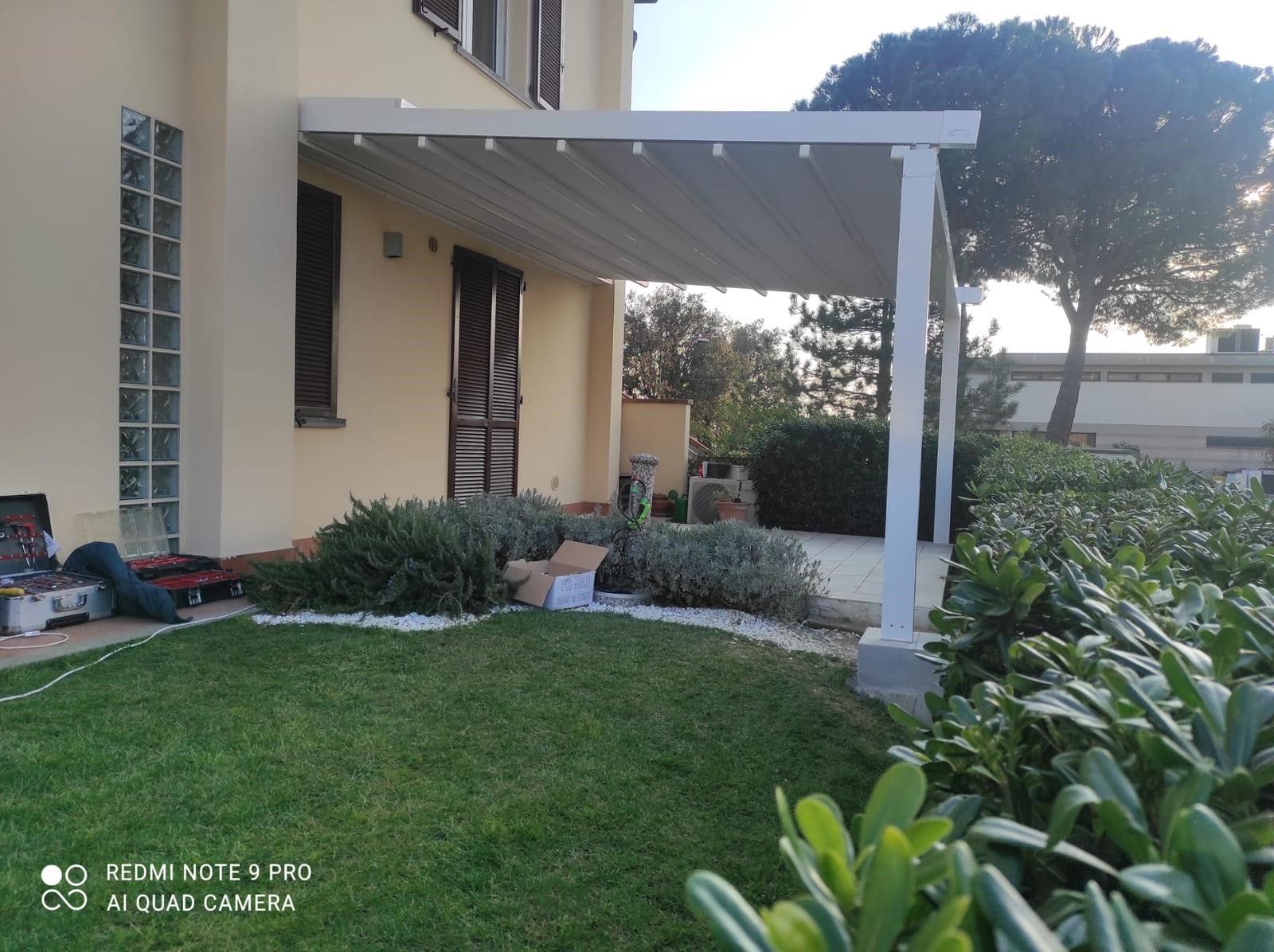 Un pergolato bianco attaccato a una casa a due piani, che fornisce ombra a un patio con verde e prato erboso.