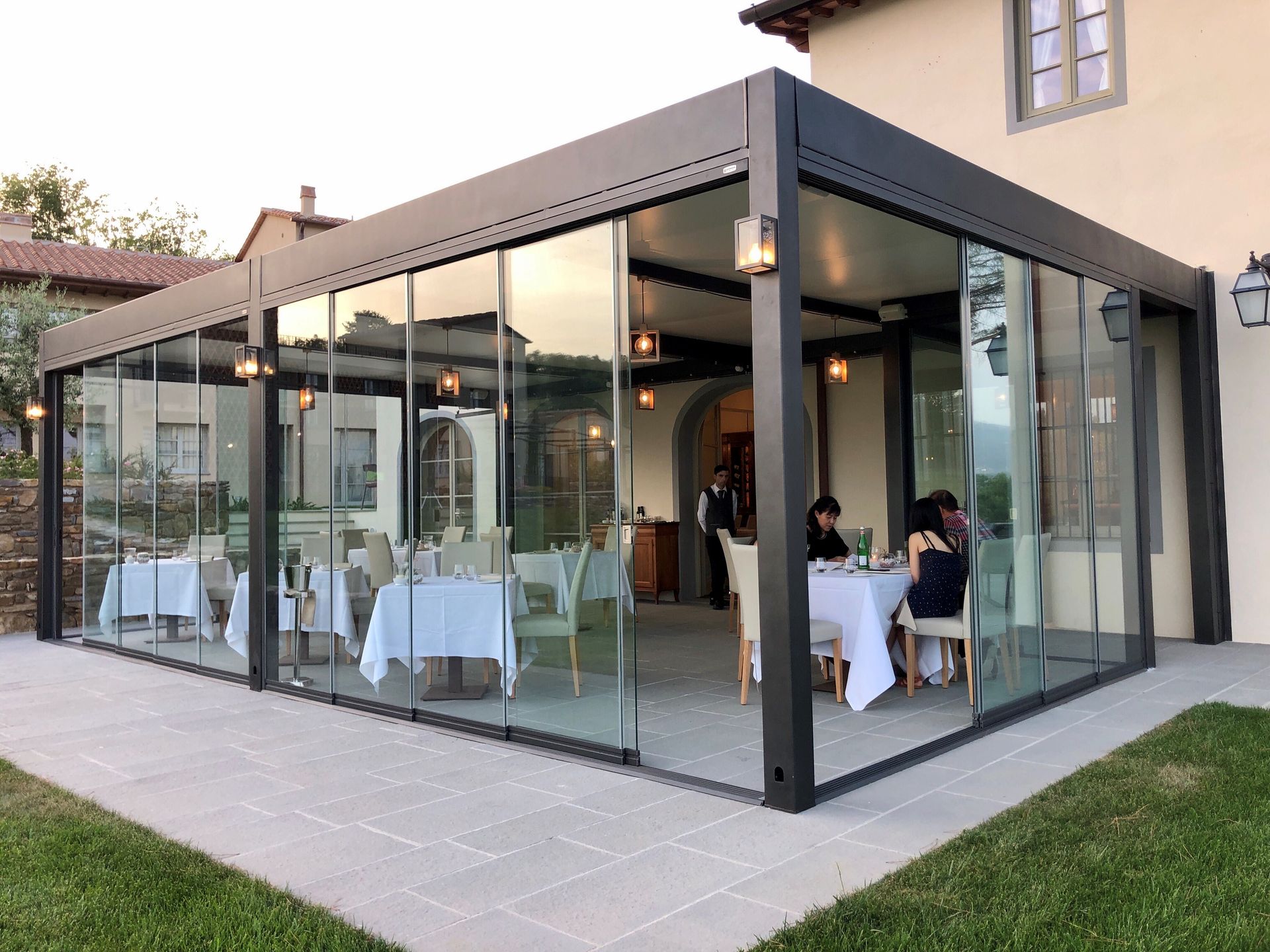 Zona pranzo esterna con vetrate e tavoli apparecchiati per i pasti, annessa a un edificio con prato erboso.