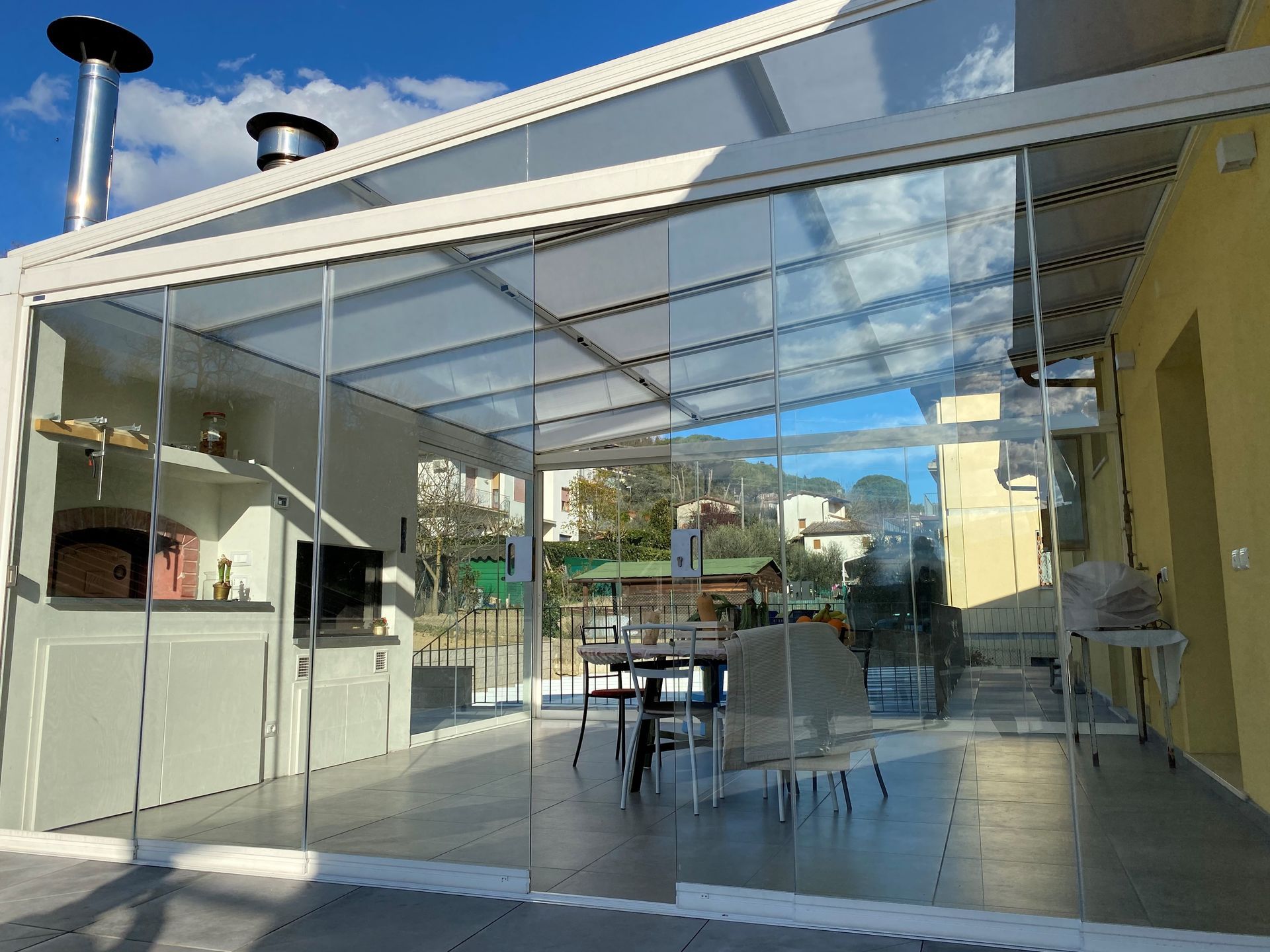 Cucina esterna con pareti in vetro, forno, tavolo e sedie, pavimenti grigi e cielo soleggiato.