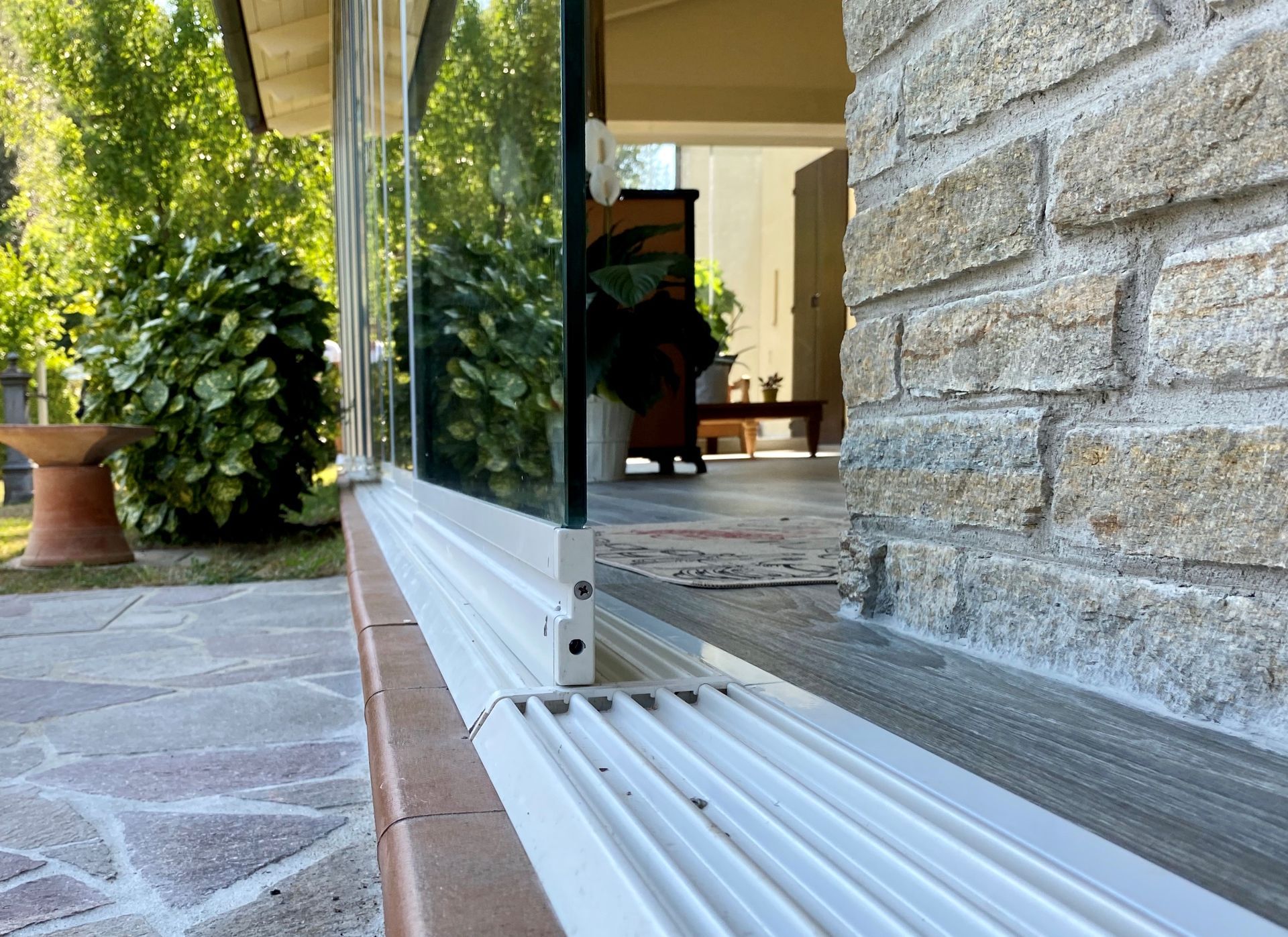 Vista dal basso di una porta scorrevole in vetro parzialmente aperta accanto a un muro di mattoni. Ambiente esterno con pietre e piante da patio.