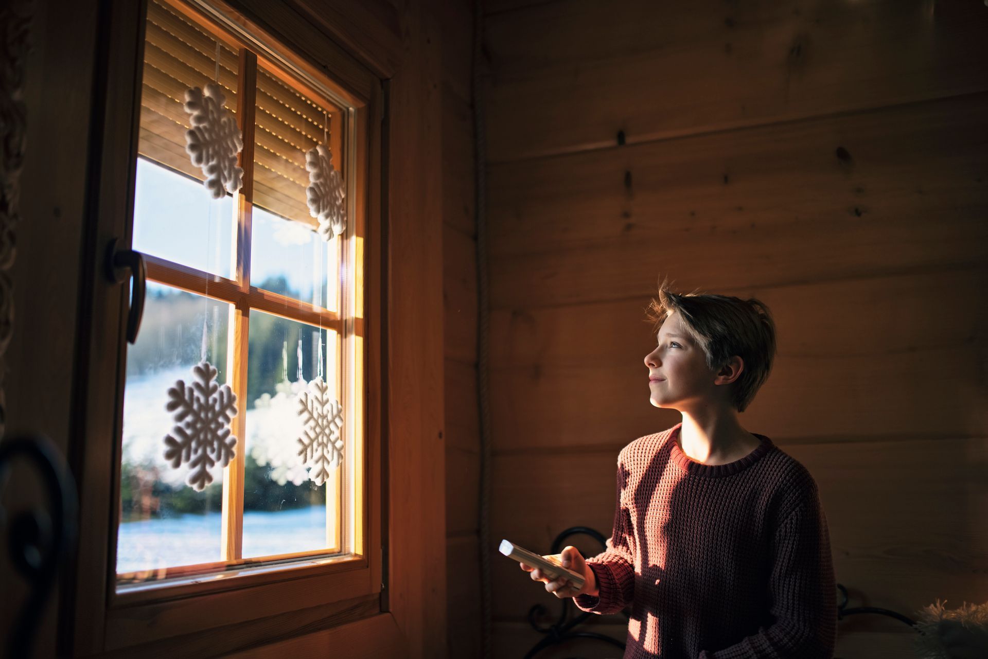 Persona che guarda fuori dalla finestra, tenendo in mano un dispositivo. Paesaggio innevato visibile. Luce calda sulle pareti interne in legno.