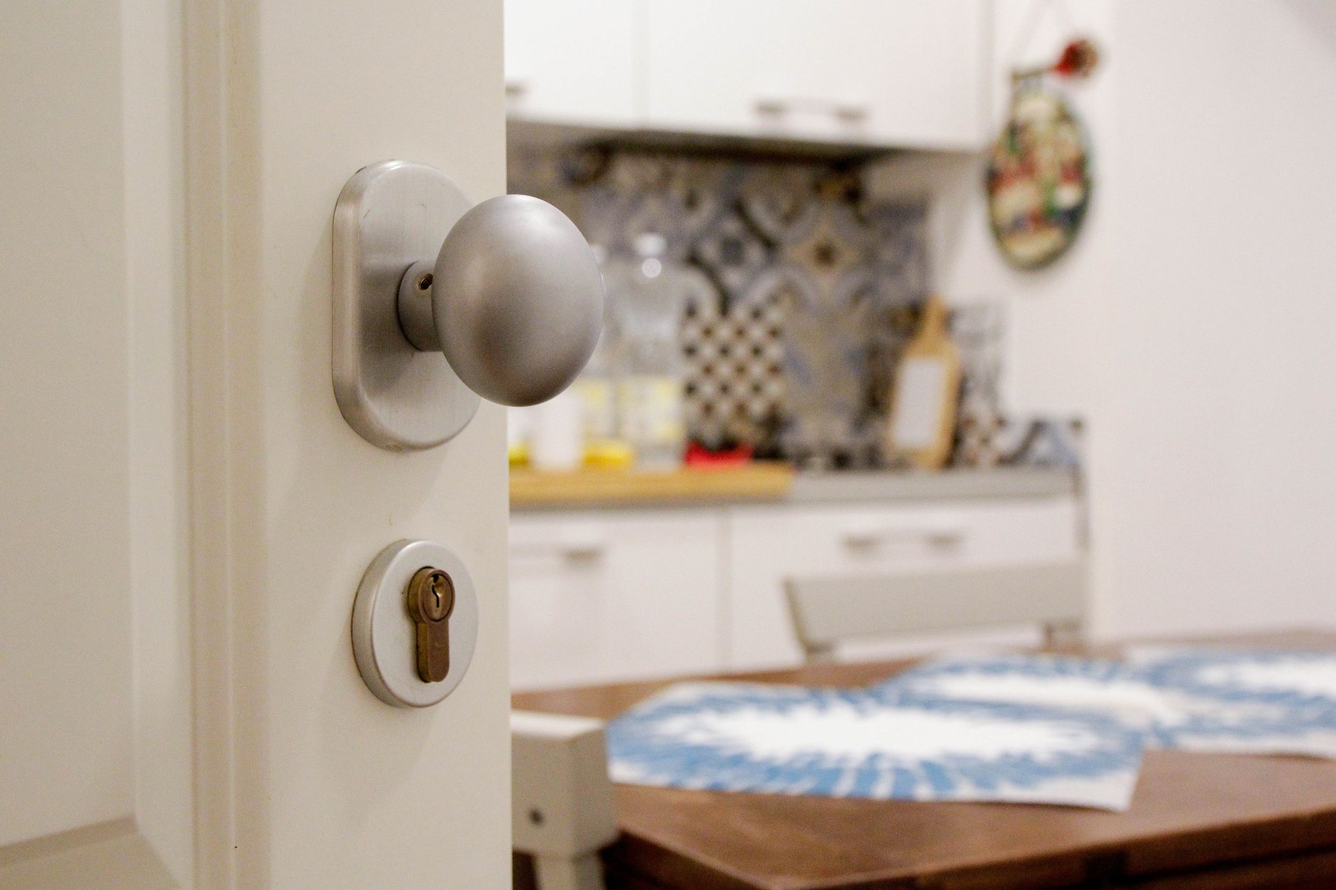 Porta socchiusa che rivela una cucina con tavolo e ripiani; maniglia e serratura visibili in primo piano.