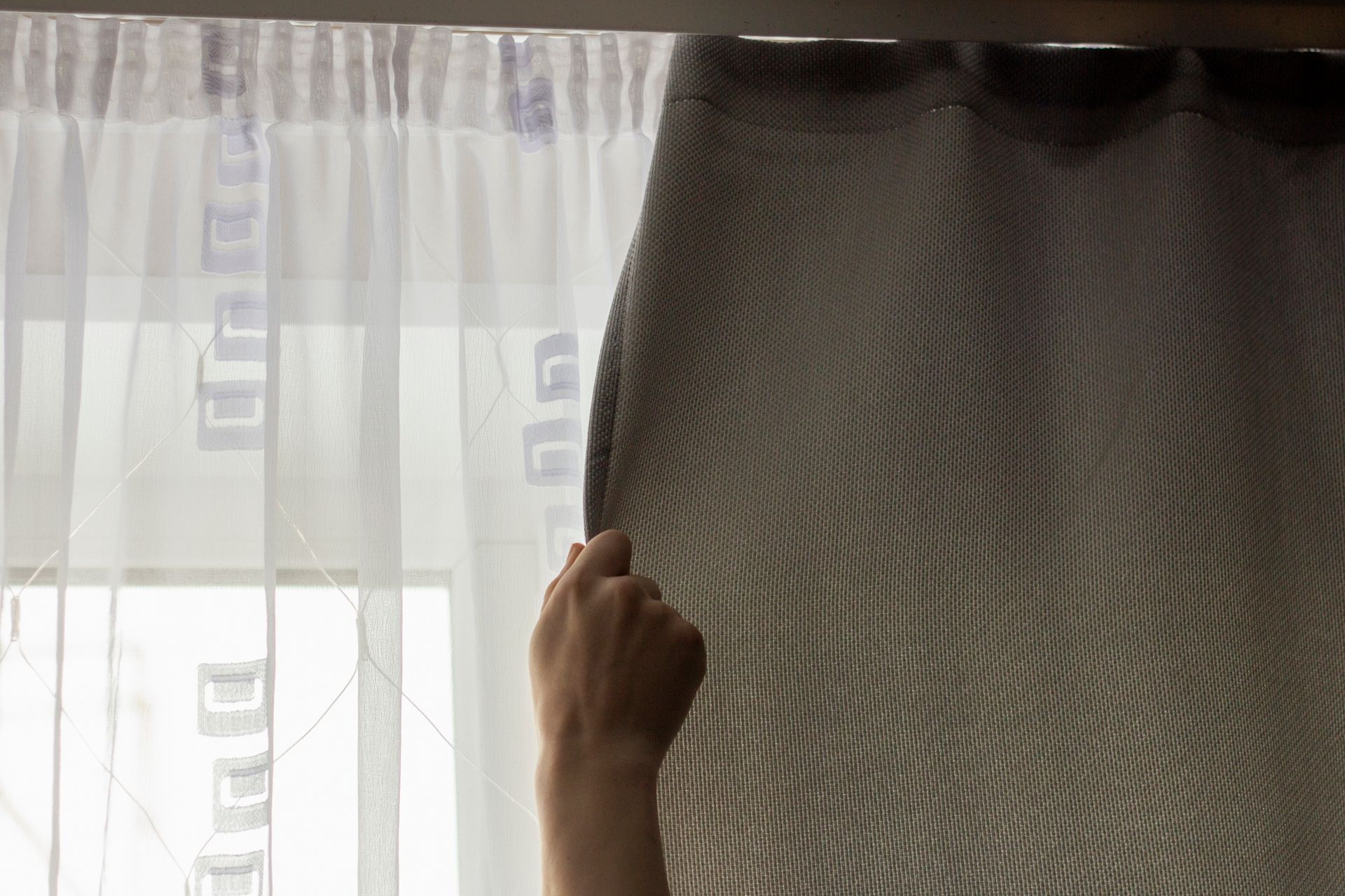 Una mano scosta una tenda grigia e rivela una finestra con una tenda bianca trasparente.