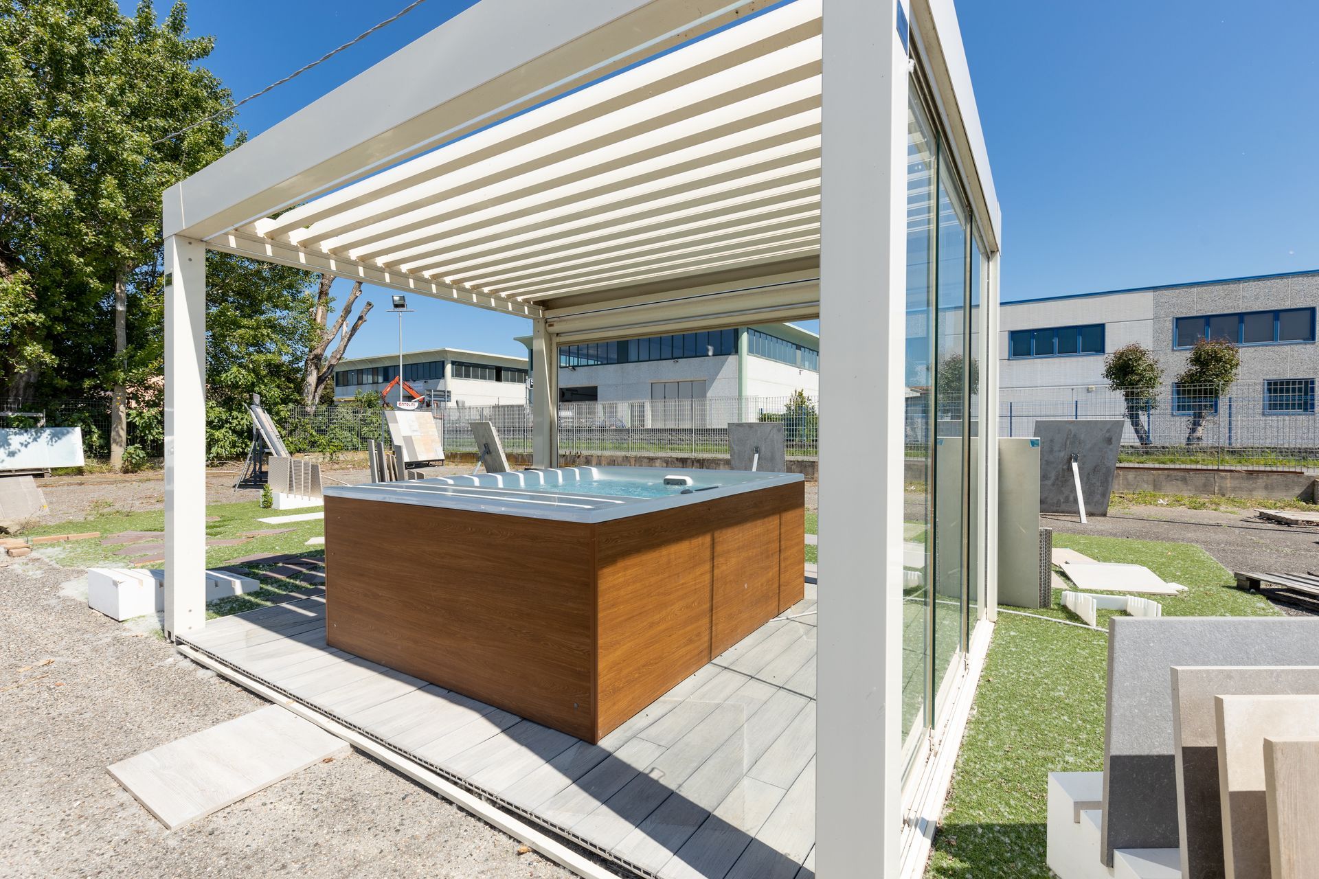 Pergolato con vasca idromassaggio, pannelli in legno, parete in vetro e tetto a doghe. Esterno, colori chiari, soleggiato.