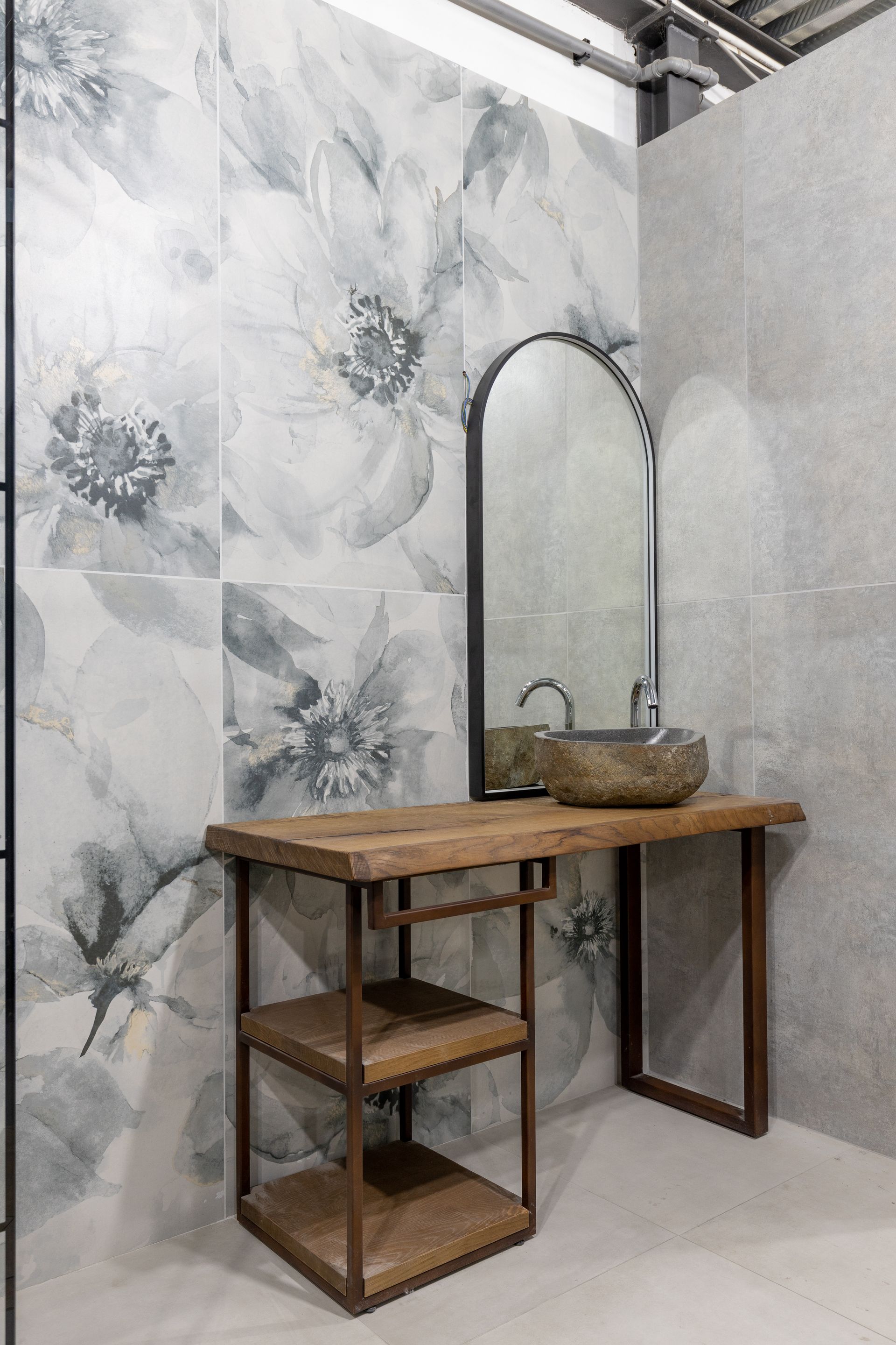 Bagno con parete piastrellata con motivi floreali, mobile lavabo in legno con mensola, lavandino in pietra, specchio ad arco.