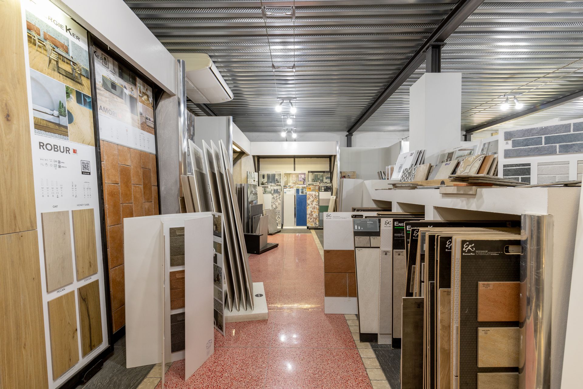 Interno di uno showroom di piastrelle con vari campioni esposti su scaffali e pareti. Pavimento a macchie rosse e bianche.