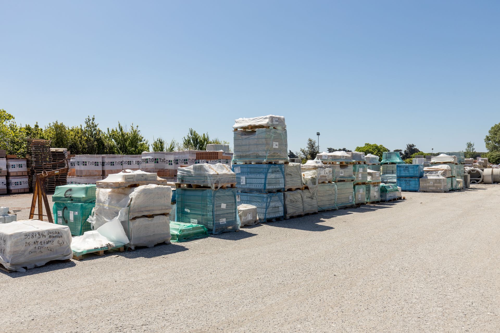 Deposito all'aperto con pile di materiali edili, pallet e sacchi sotto un cielo azzurro e limpido.