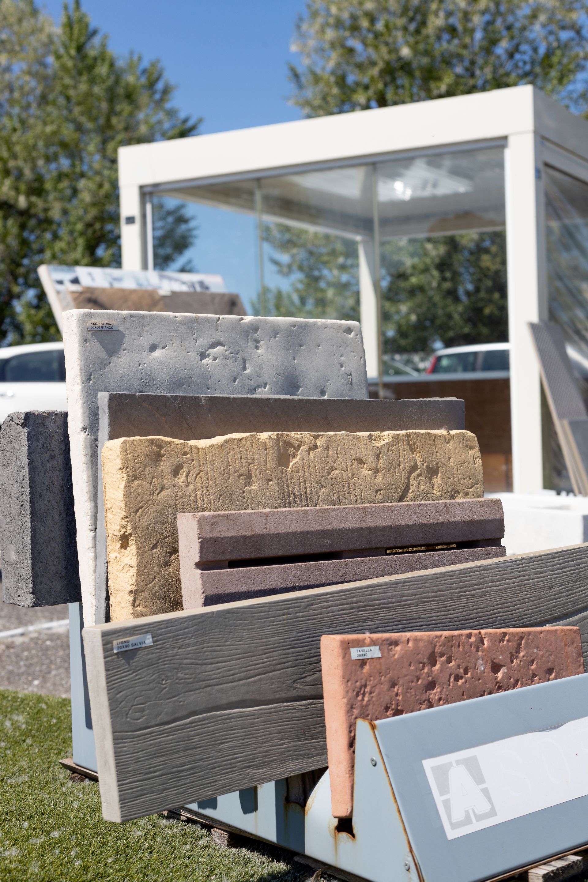 Esposizione di piastrelle in pietra di vari colori all'aperto, con una struttura bianca sullo sfondo.