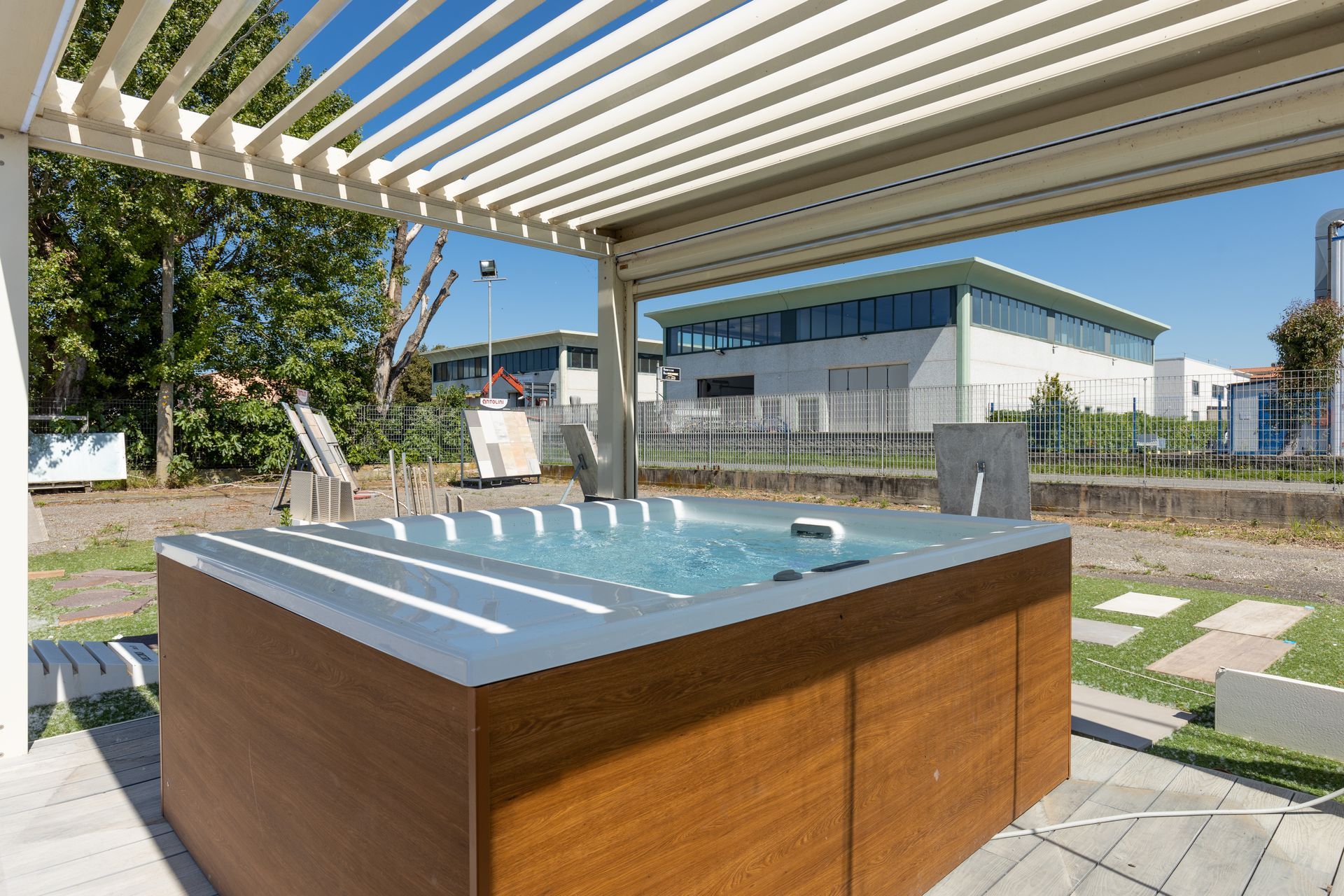 Vasca idromassaggio sotto un pergolato in un ambiente esterno. Pannelli in legno, acqua azzurra e pavimentazione grigia.