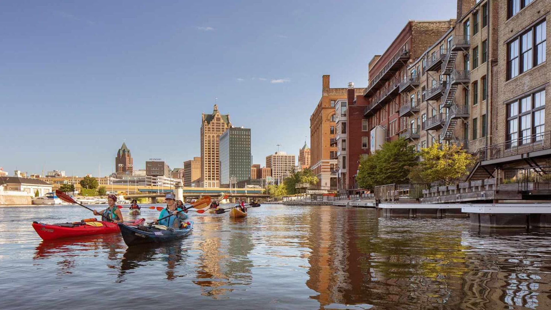 Explore the scenic Milwaukee RiverWalk filled with activities and waterfront dining. View of Milwaukee RiverWalk with kayakers and boats on a sunny summer day.