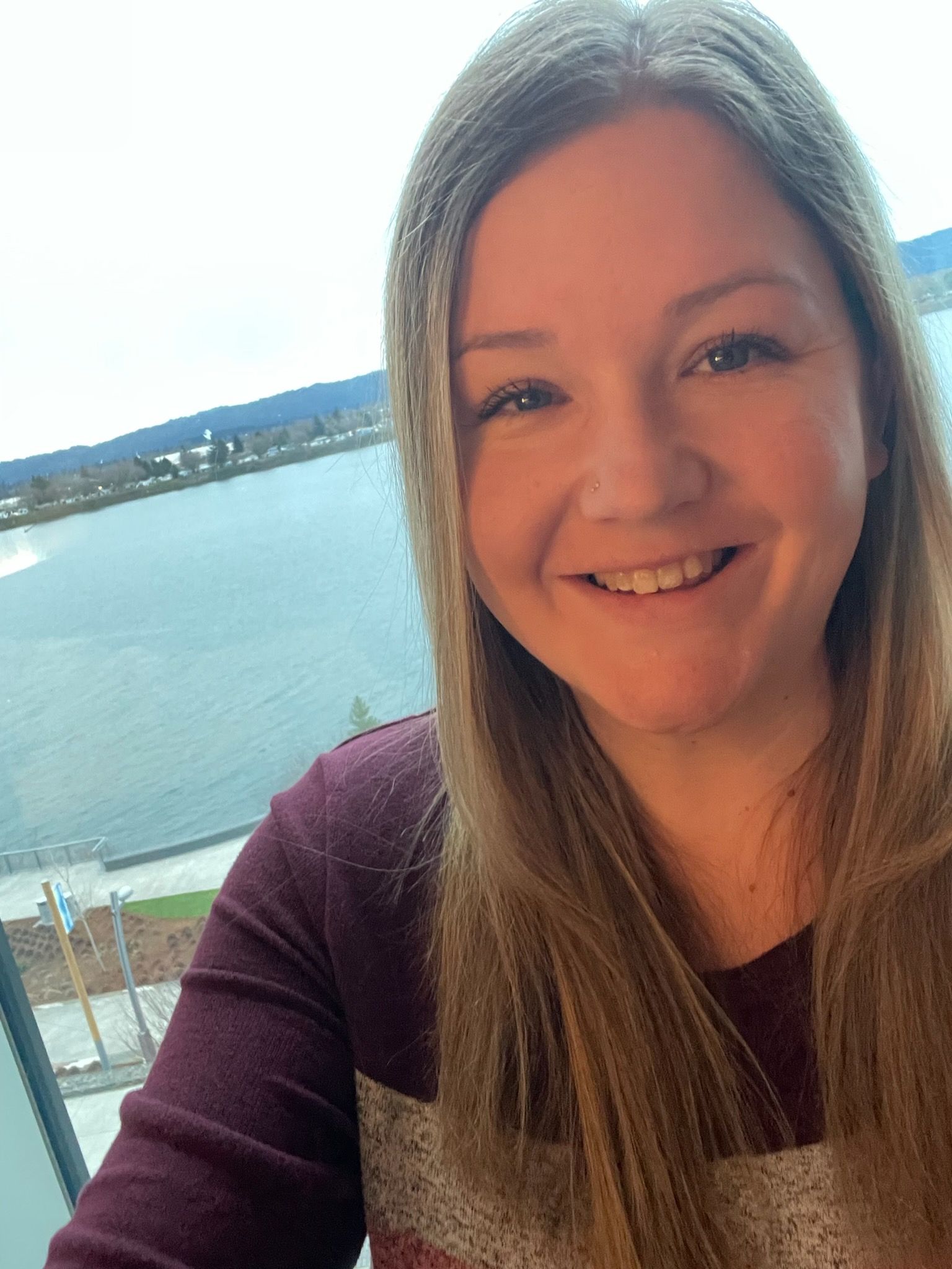 A woman is smiling in front of a window overlooking a body of water.