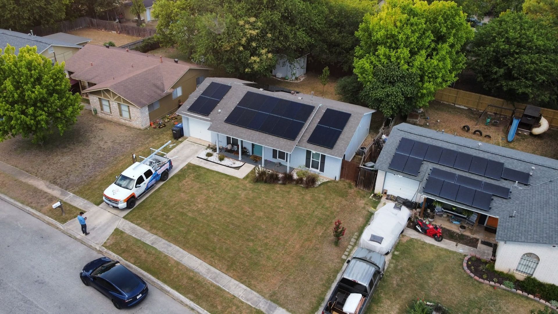 Aerial view of suburban homes with solar panels, parked vehicles, and a grassy yard.