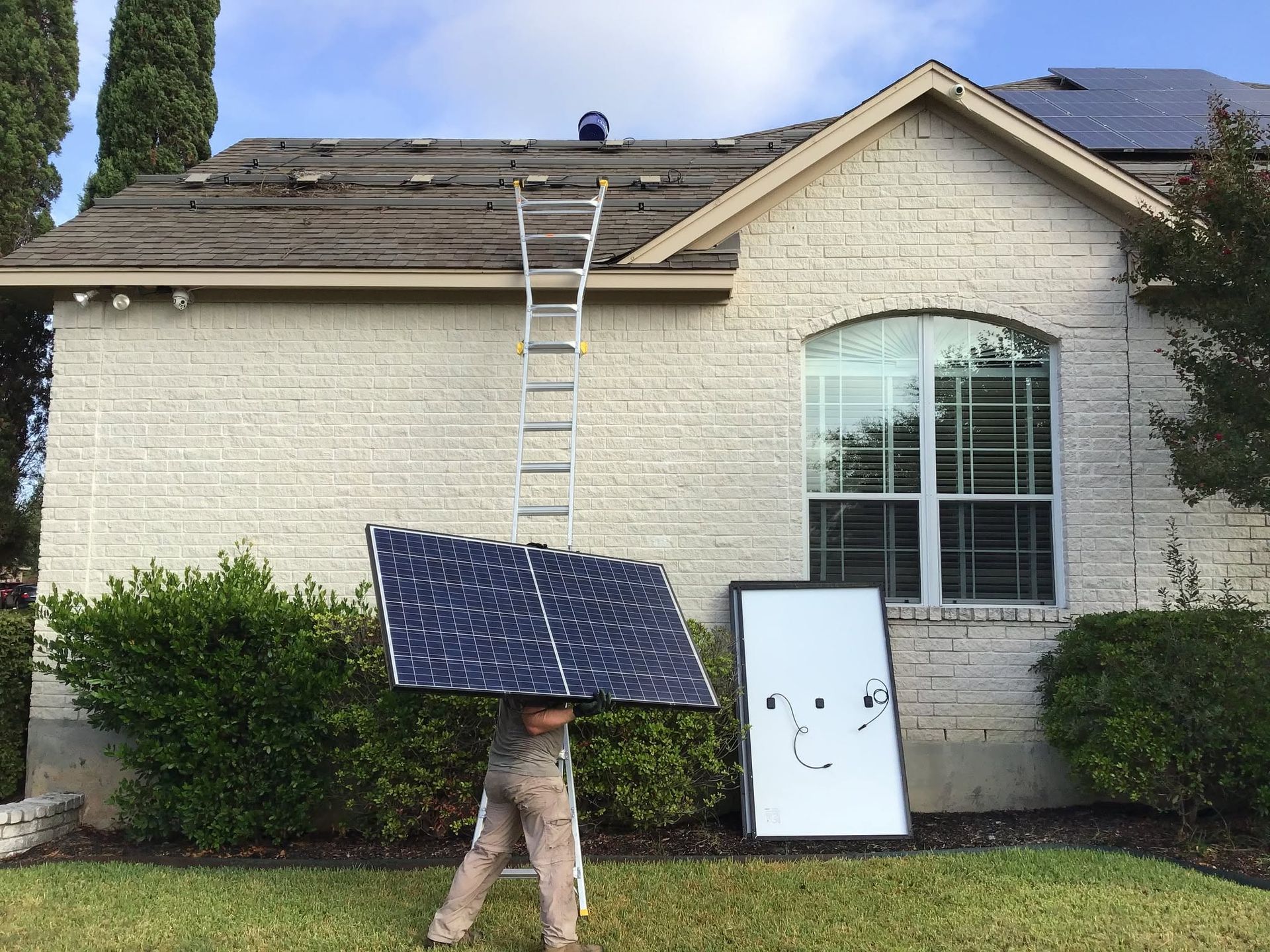 Person holding a solar panel on a ladder beside a house with a white cabinet outdoors.