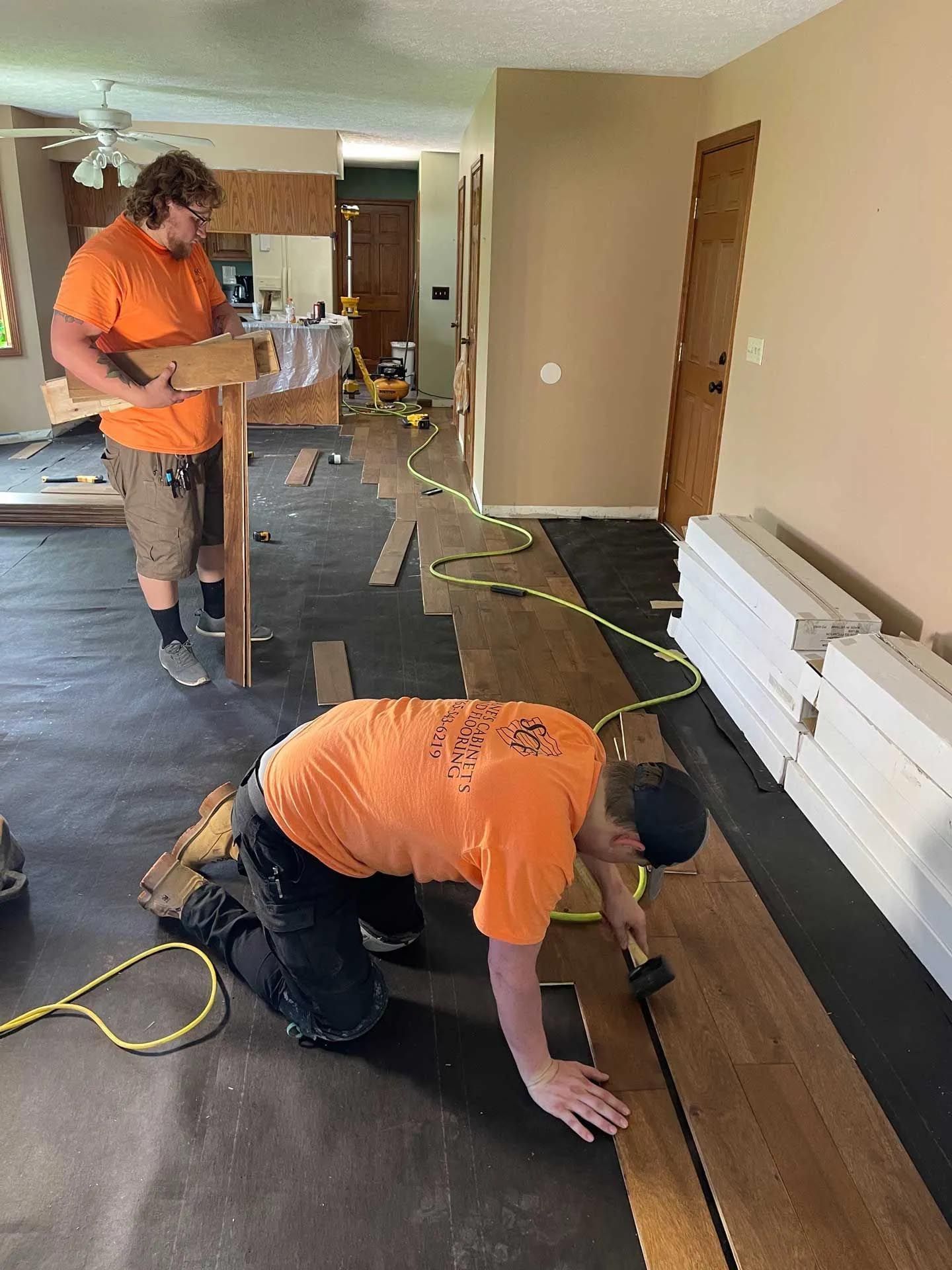 Two men are installing hardwood flooring in a living room.