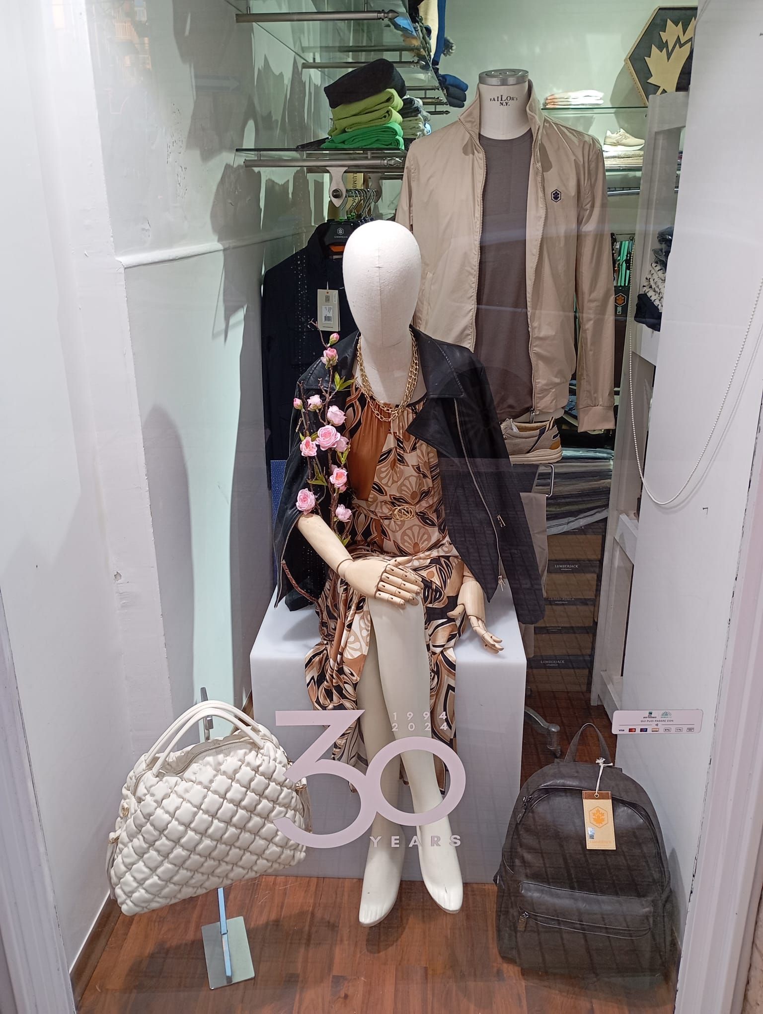 Un manichino è seduto nella vetrina di un negozio accanto a uno zaino e una borsa.