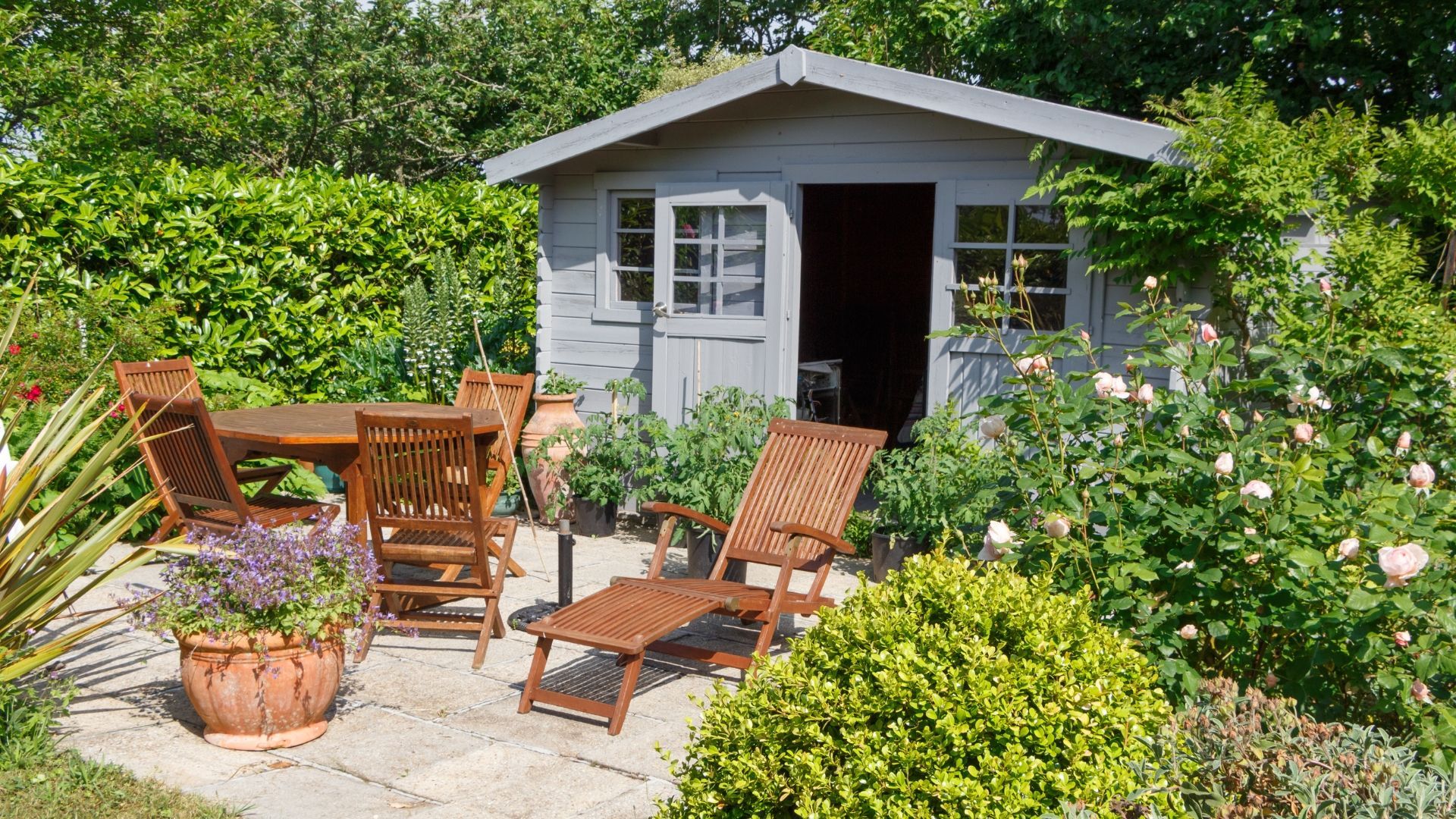 Wooden patio set next to a gray shed in a lush garden.