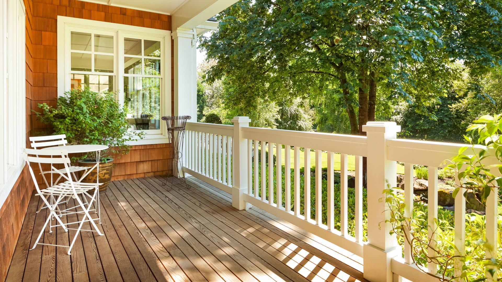 Wooden porch with white railing, table, chairs, and lush greenery in the background.