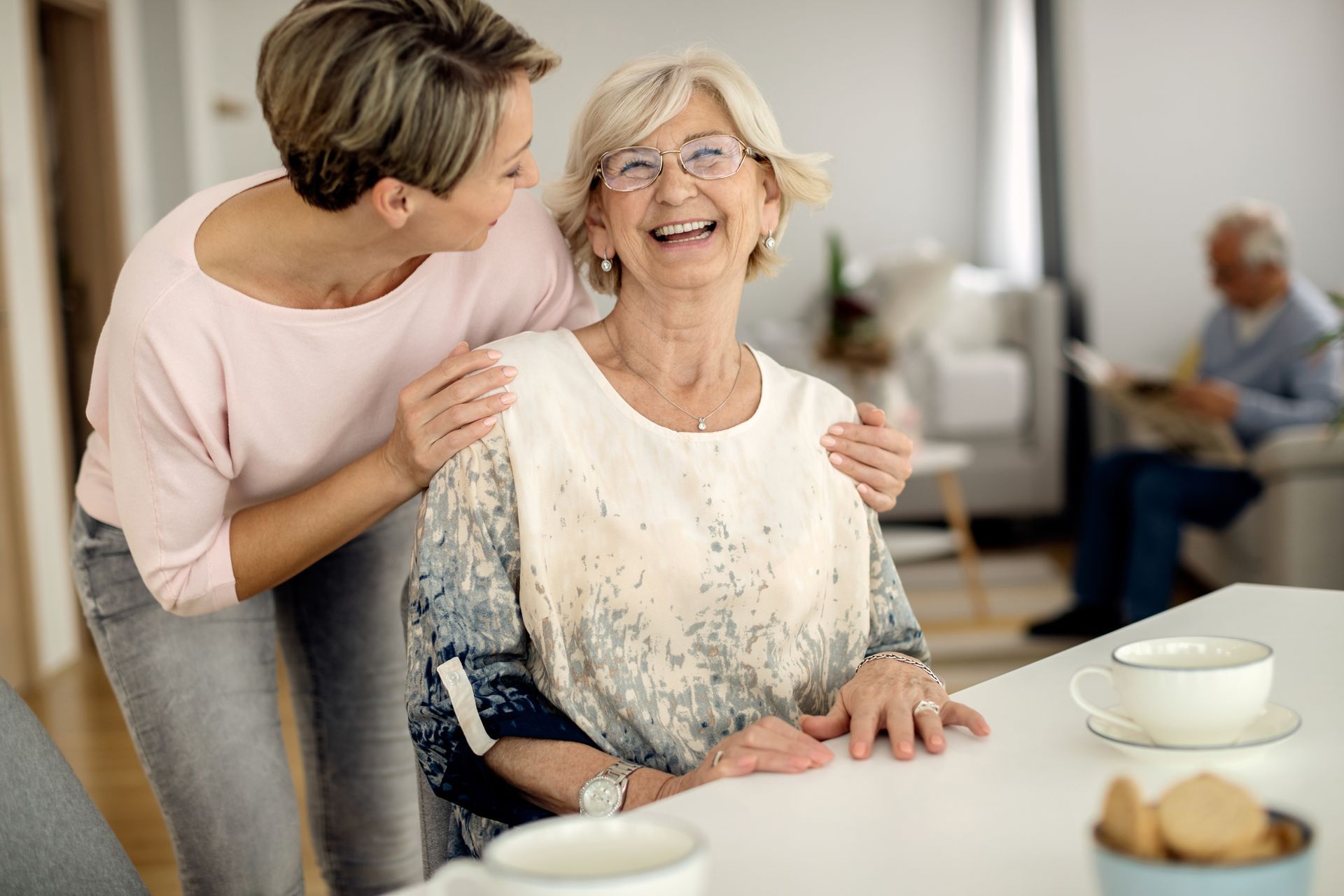 Woman hugs older woman who laughs, both indoors. Man reads in background.