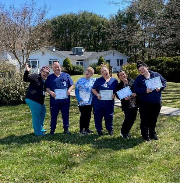 Group of seven healthcare workers outside a house, smiling, holding certificates. Sunny day.