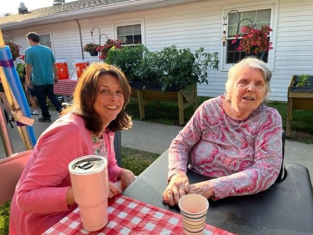 Two women smiling at a table outdoors. One in pink, the other in a patterned shirt. Garden boxes and a building are in the background.