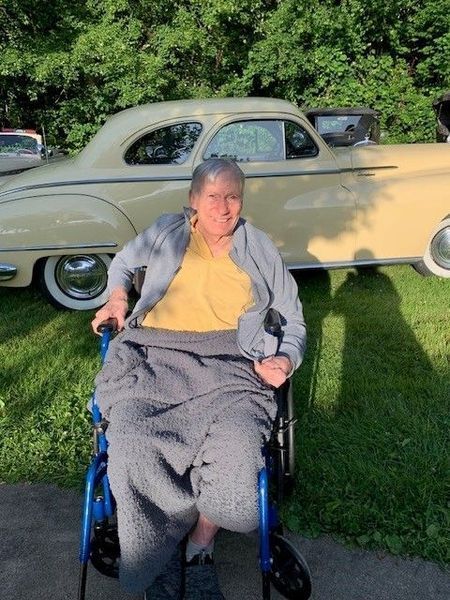 Woman in wheelchair, gray blanket, yellow shirt, in front of a classic yellow car. Outdoors, green grass.