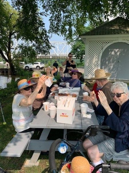 People seated at a picnic table outdoors. Some are clapping, enjoying food. A bridge is visible in the background.