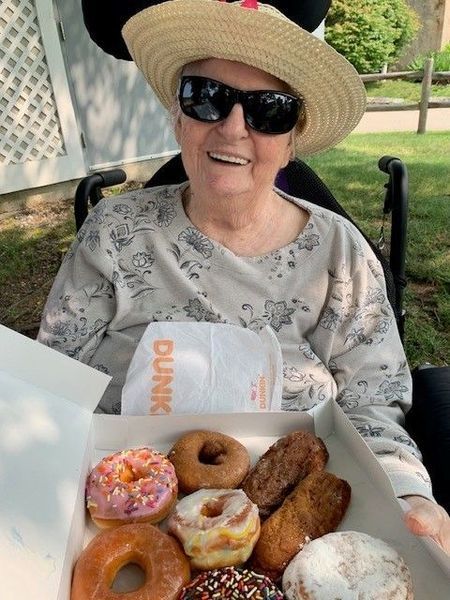 Woman in hat and sunglasses smiles while holding a box of Dunkin' Donuts outdoors.