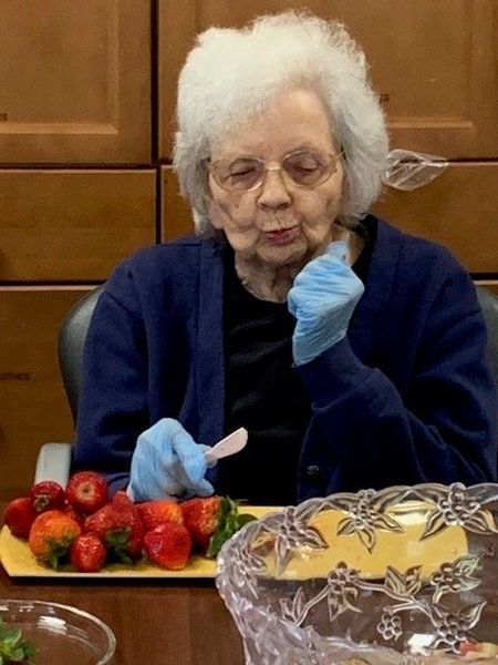 Elderly woman eating strawberries, wearing gloves, at a table.