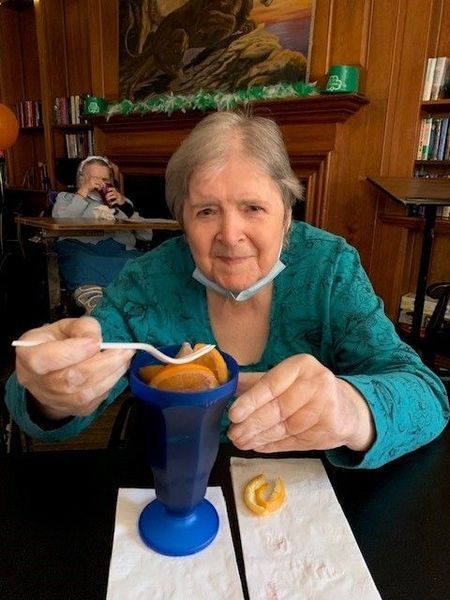 Elderly woman in teal top, smiling, eating dessert in blue cup. Table setting with napkin and fortune cookie.