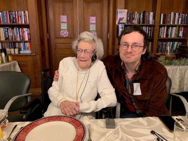 An older woman in a wheelchair with headphones and a younger man with arm around her, both smiling.