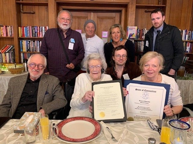 Group of people with an older woman holding framed documents, likely an award, at a table in a library.