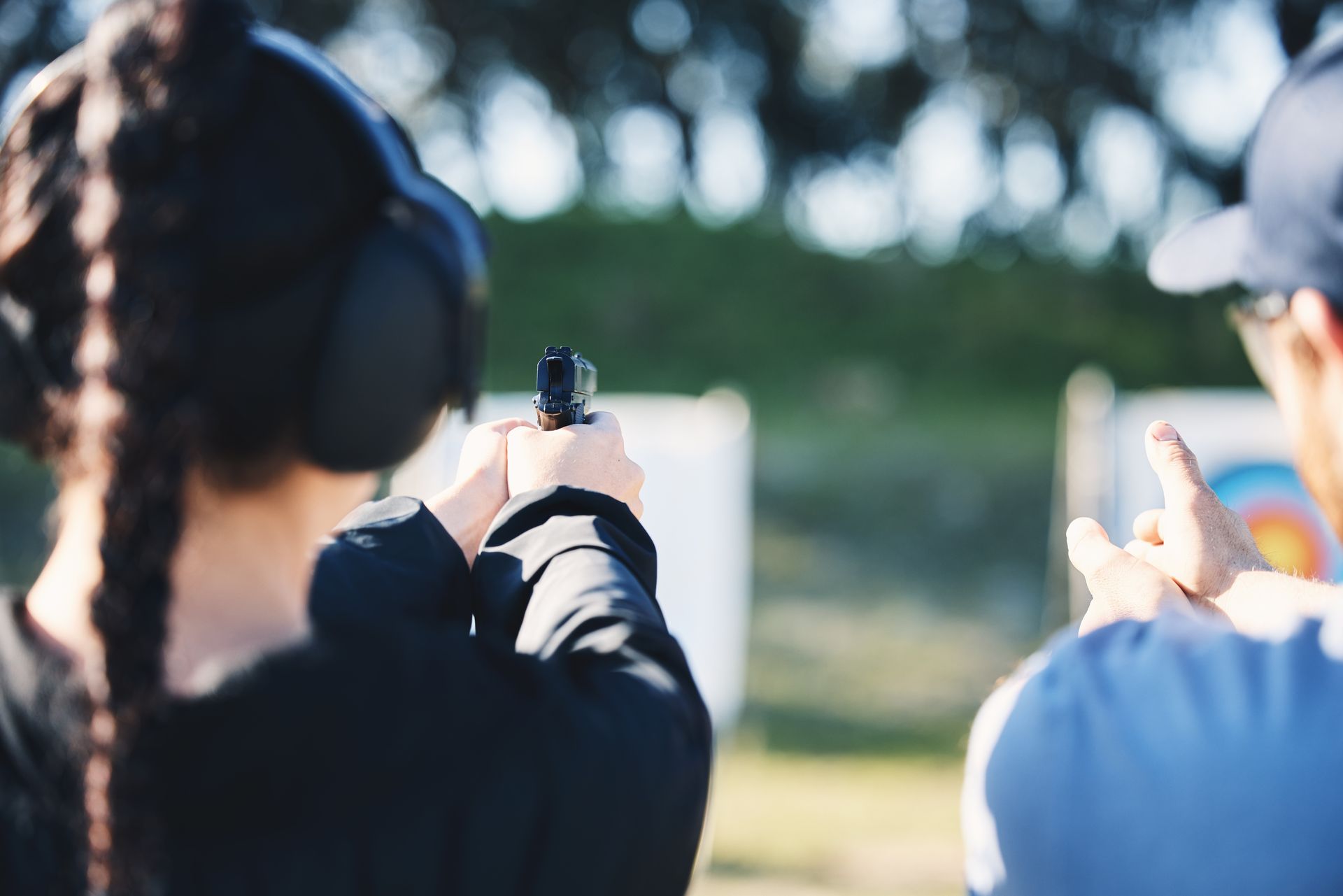 Person aiming a handgun at a target under the supervision of another person at an outdoor shooting range.