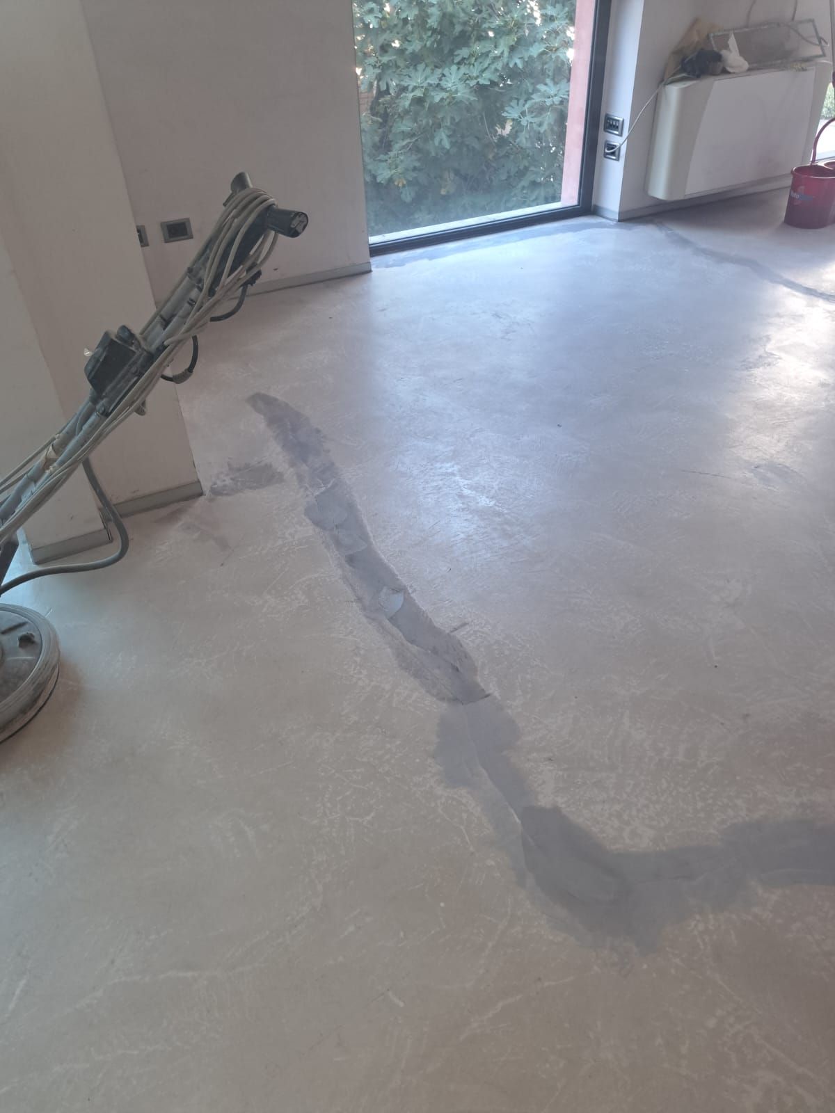 Levigatura del pavimento in corso; pavimento in cemento grigio, macchina, stanza soleggiata con finestra.