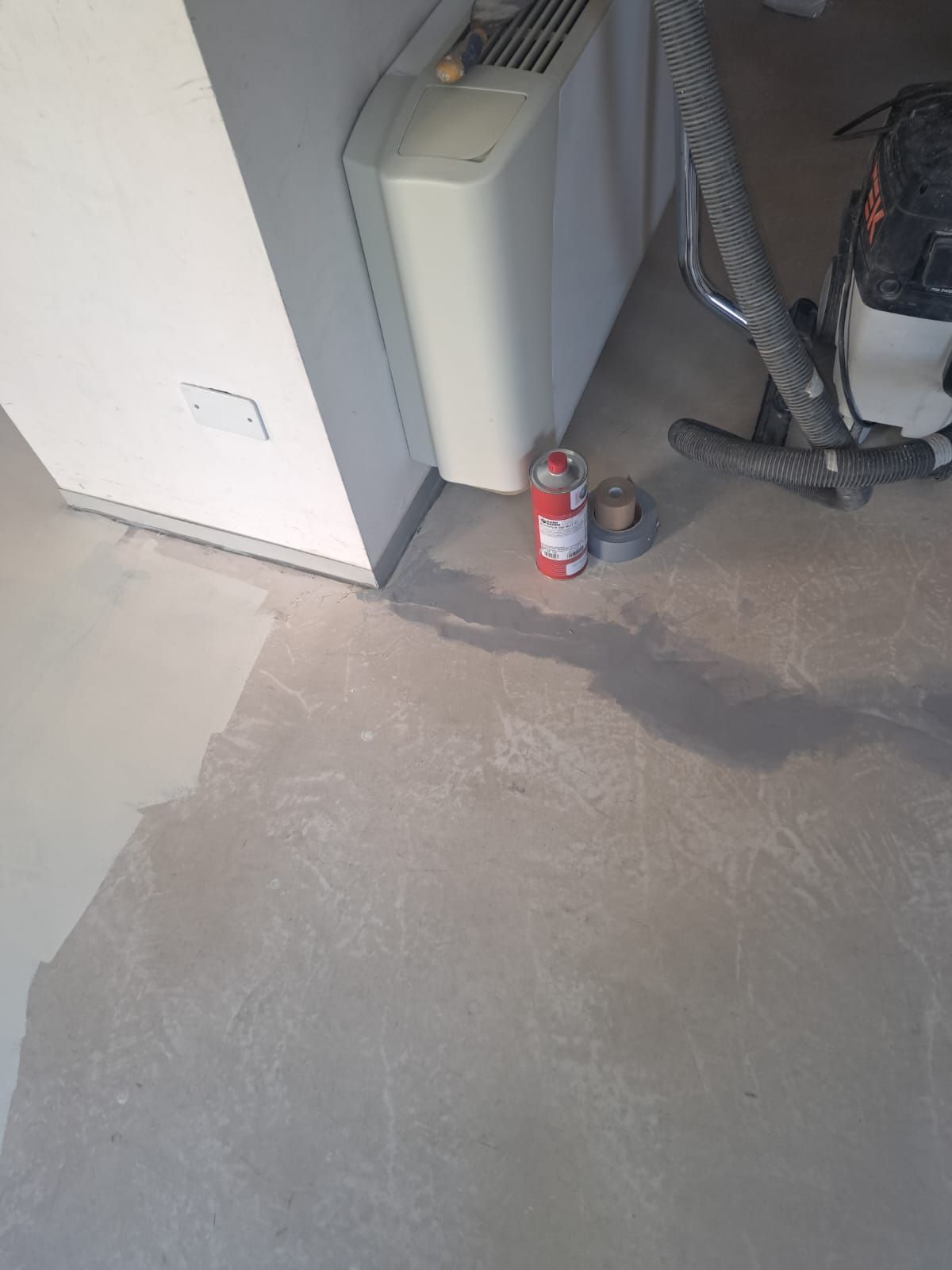 Pavimento in cemento grigio chiaro con parete grigio chiaro, un termosifone, un estintore e un aspirapolvere.