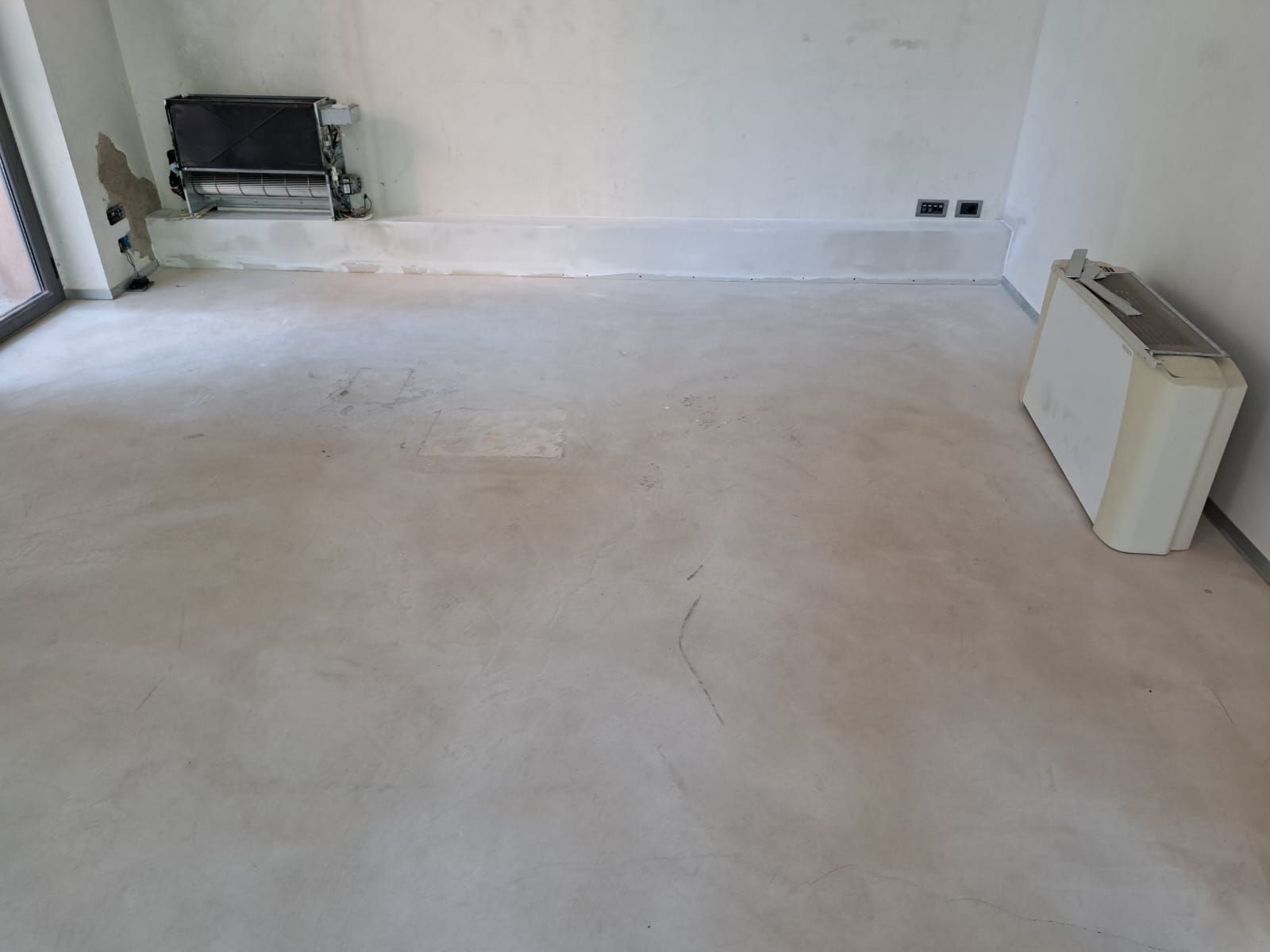 Stanza vuota con pavimento in cemento chiaro e pareti bianche; nello spazio sono presenti un termosifone e una stufa.