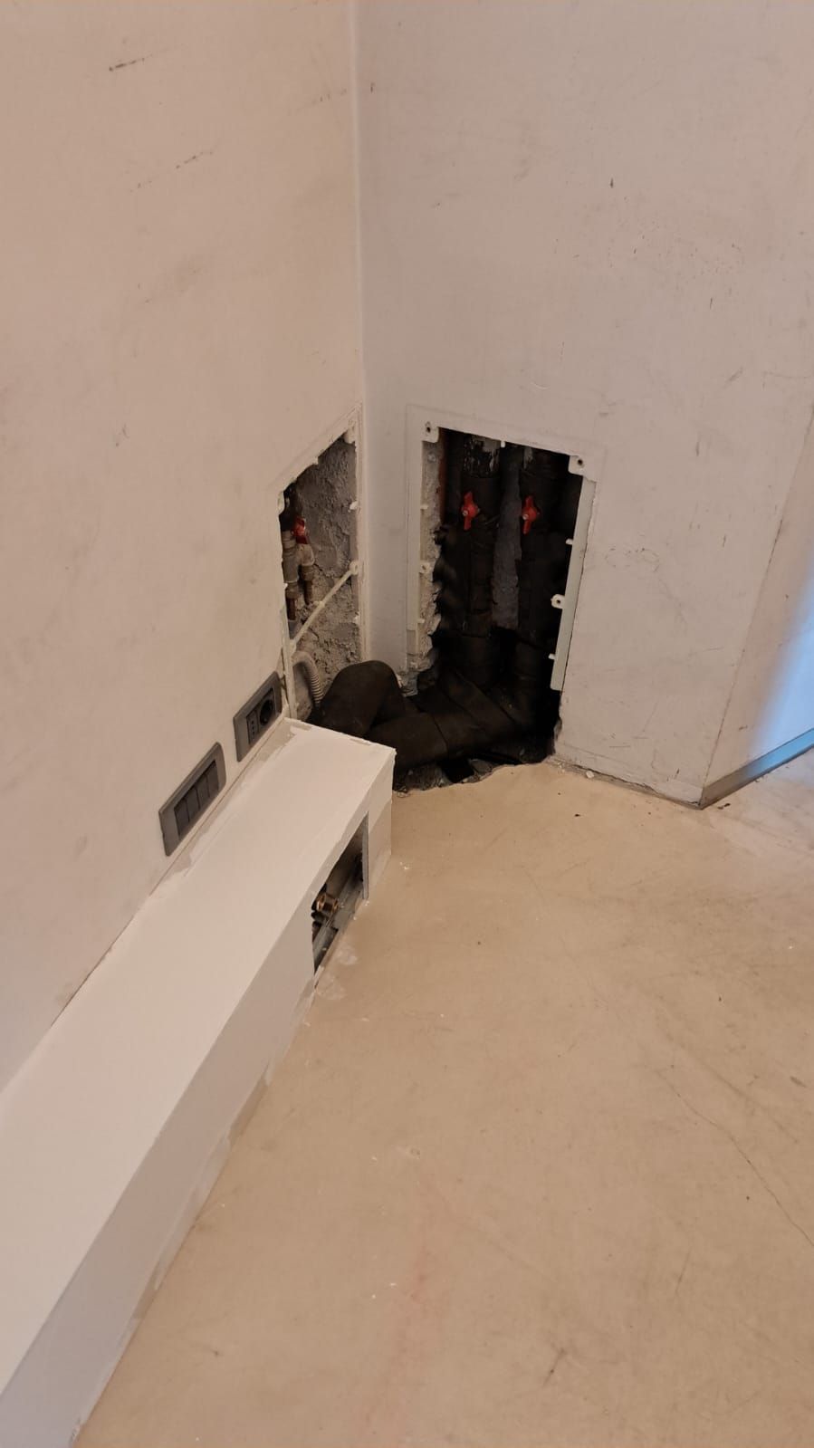 Angolo interno di una stanza in costruzione con punti di accesso all'impianto elettrico e idraulico. Pareti bianche, pavimento grigio, tubature nere.