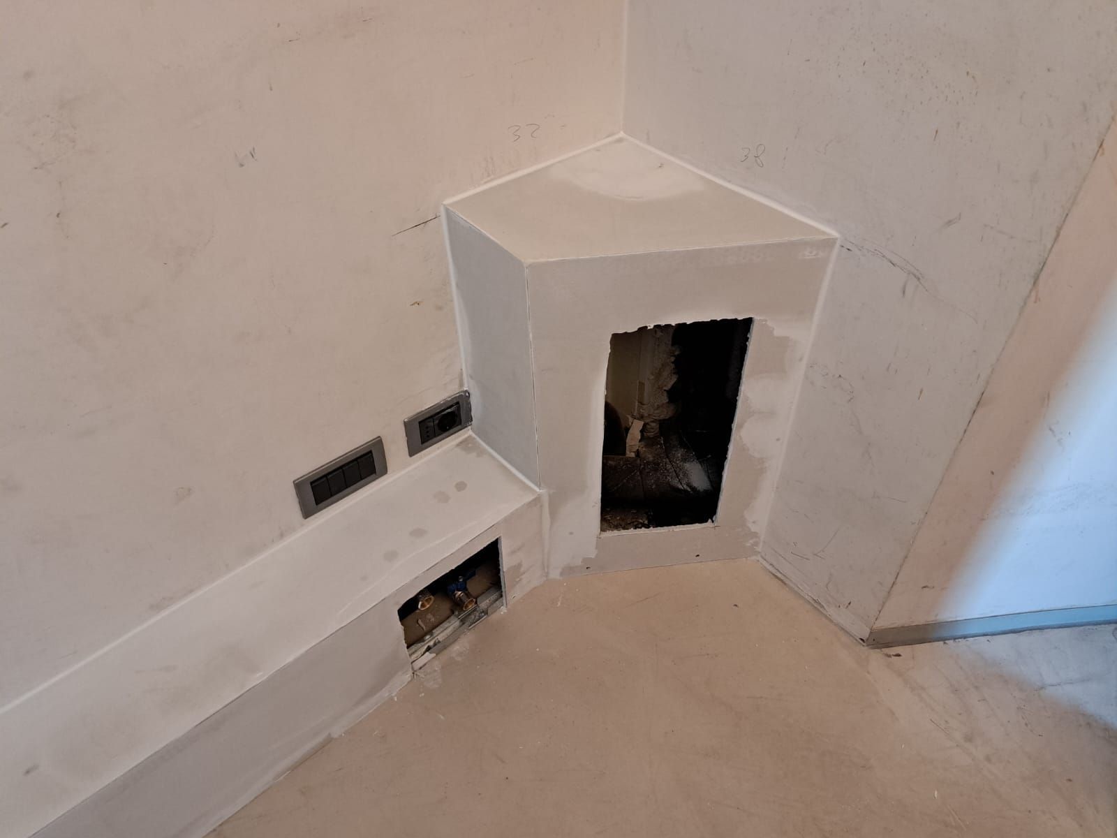 Un camino angolare in costruzione, circondato da pareti e pavimenti grezzi; include aperture di ventilazione.