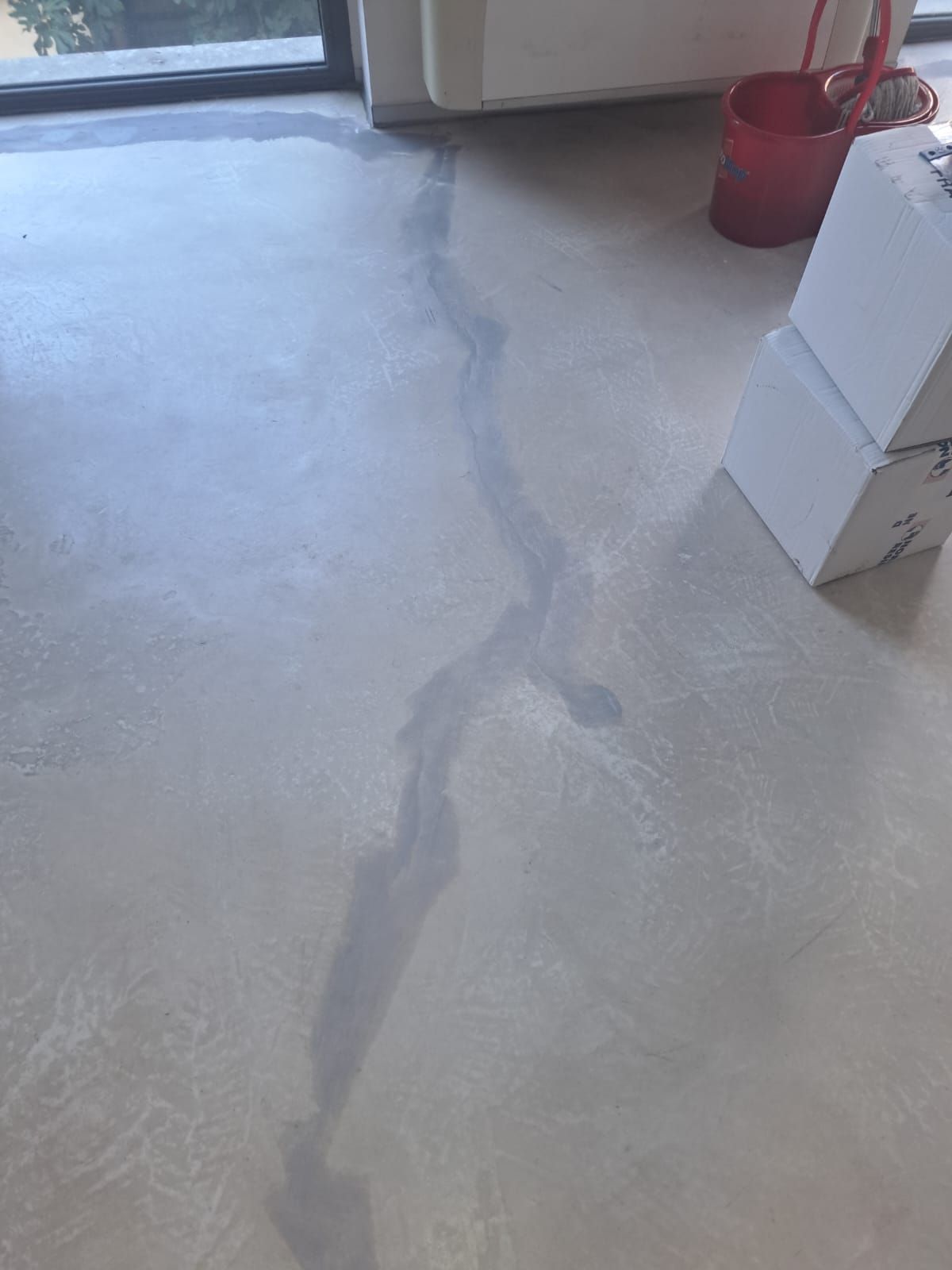 Pavimento in cemento grigio chiaro con una macchia scura bagnata, una finestra e un secchio rosso.