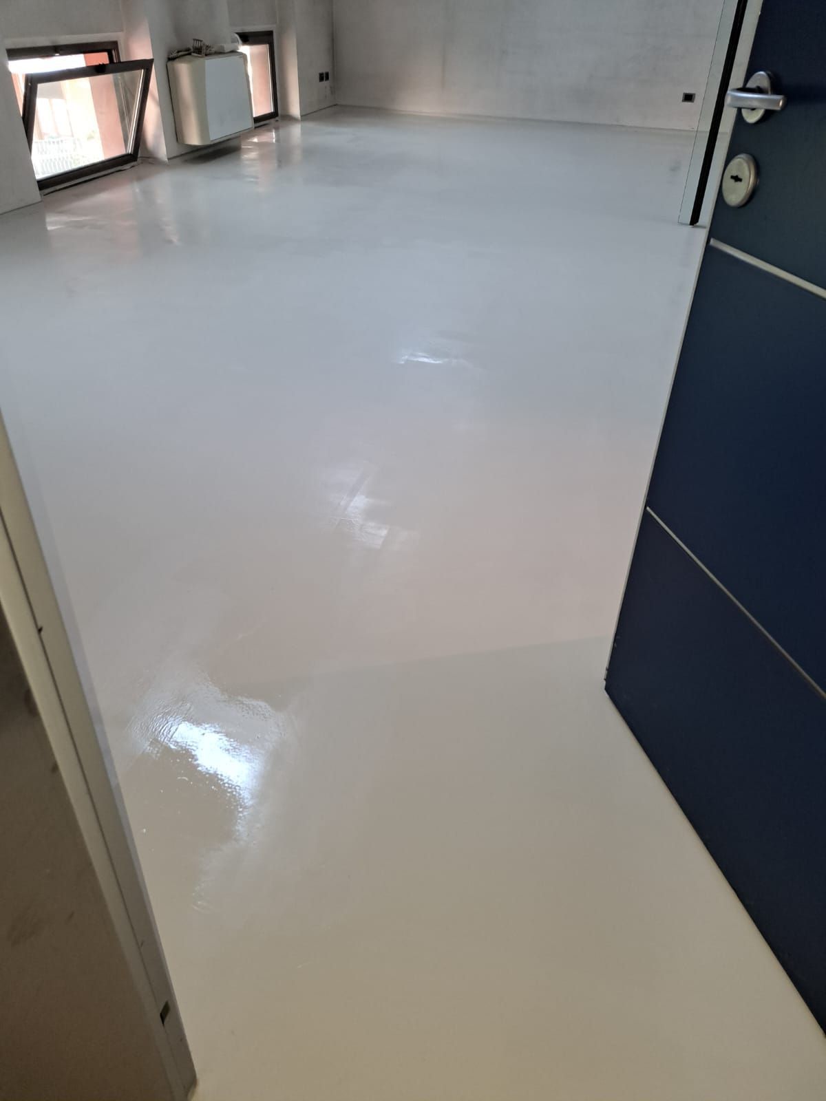 Pavimento in resina epossidica bianca in una stanza vuota, riflessi della luce solare, porta blu scuro sulla destra.