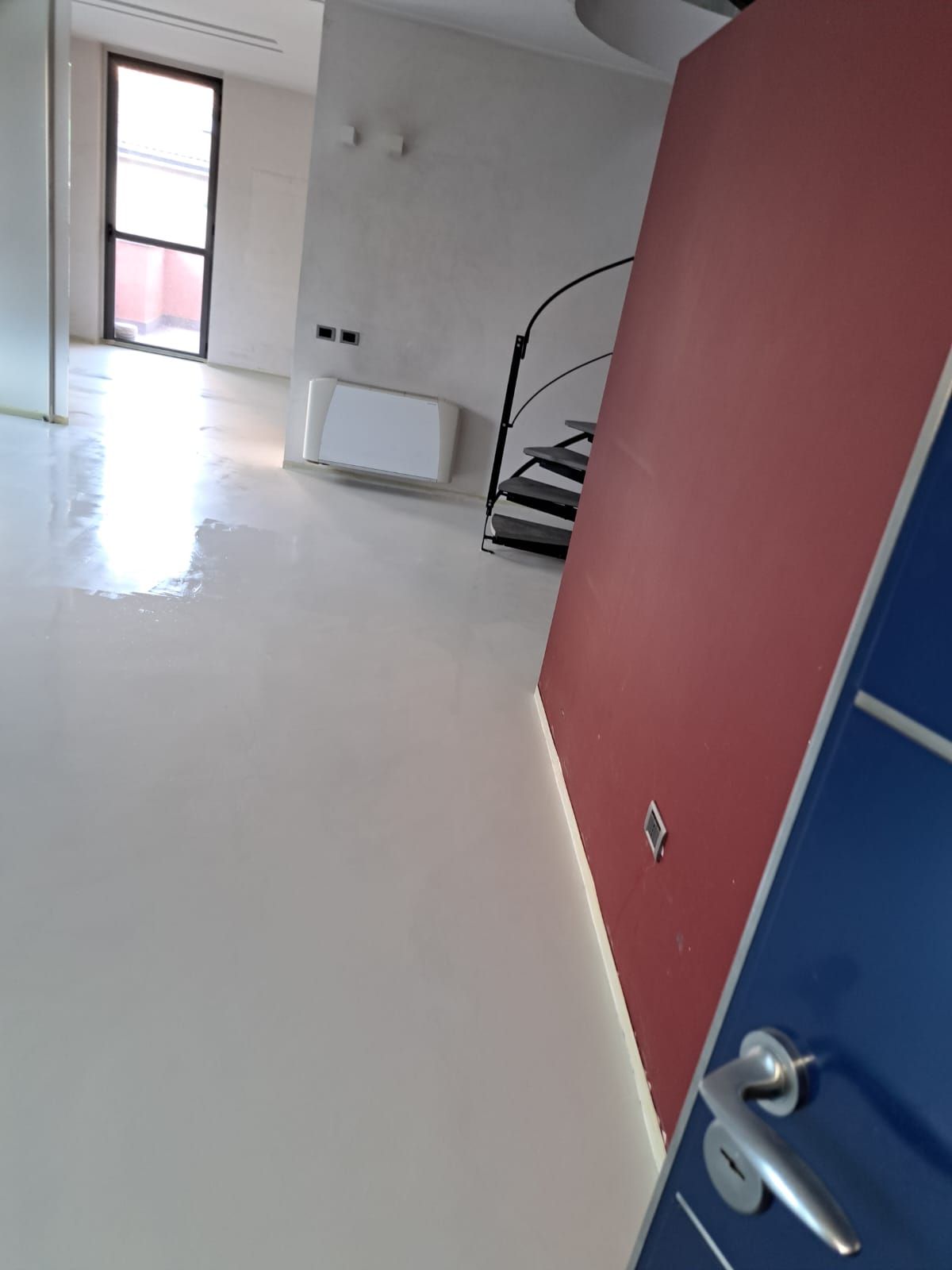 Stanza bianca con pannello rosso, porta blu, scale a chiocciola nere e finestra.