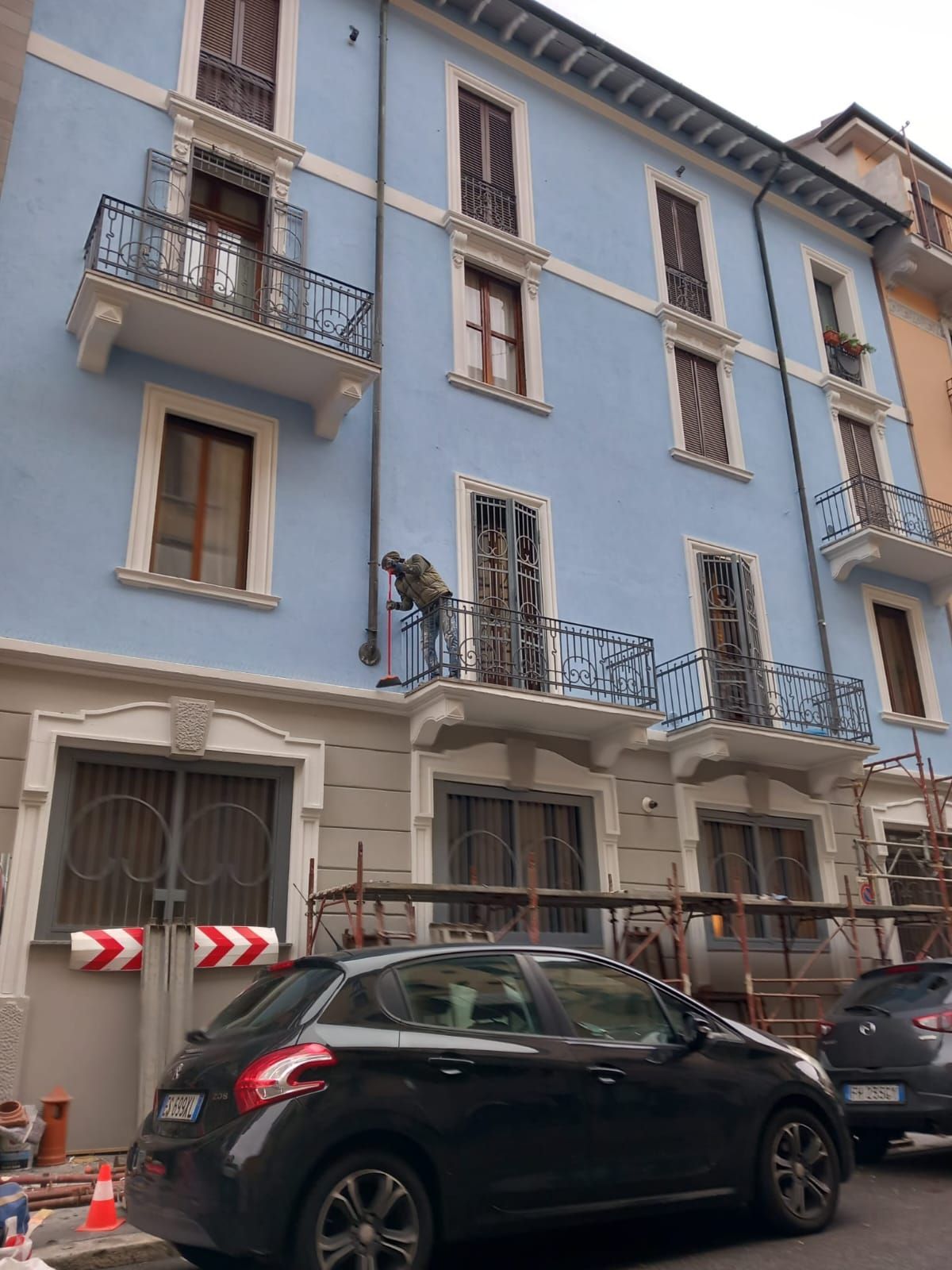 Edificio blu con balconi, persiane marroni, macchina nera parcheggiata di fronte, lavori in corso.