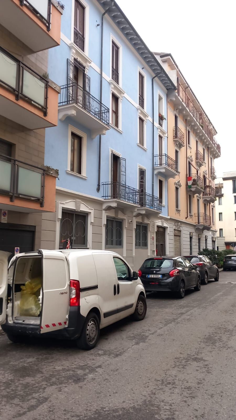 Un condominio blu e color pesca con balconi, auto parcheggiate e un furgone bianco sulla strada.