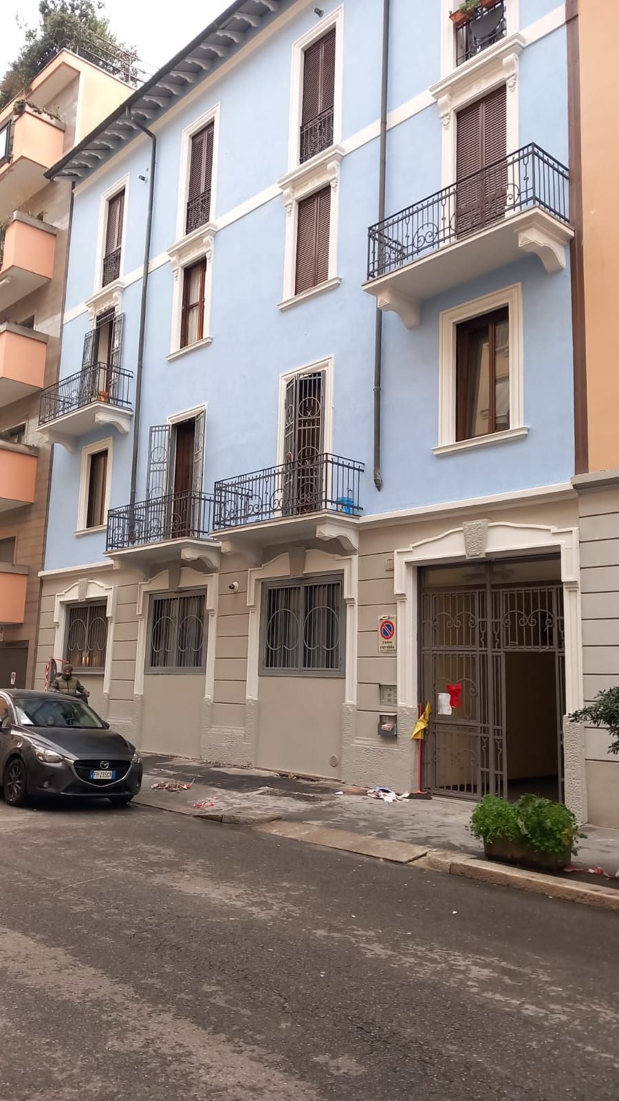 Un edificio azzurro con balconi, vista sulla strada, un'auto nera parcheggiata e un cancello d'ingresso.