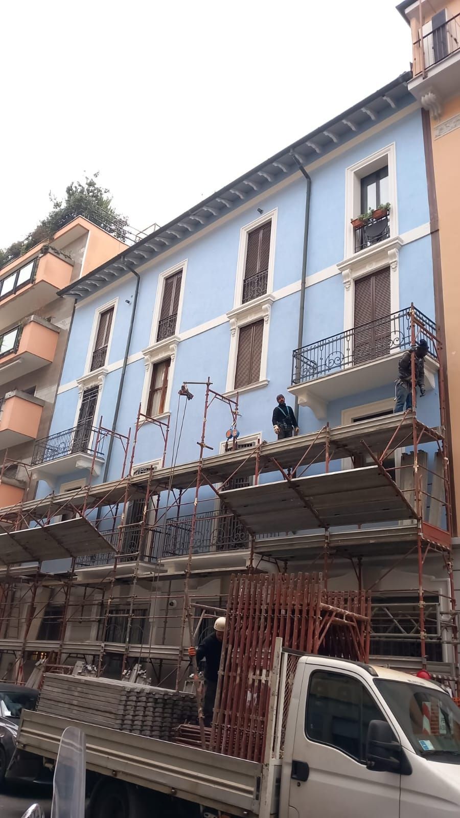 Edificio con impalcature e operai; facciata azzurra, porte/finestre marroni.