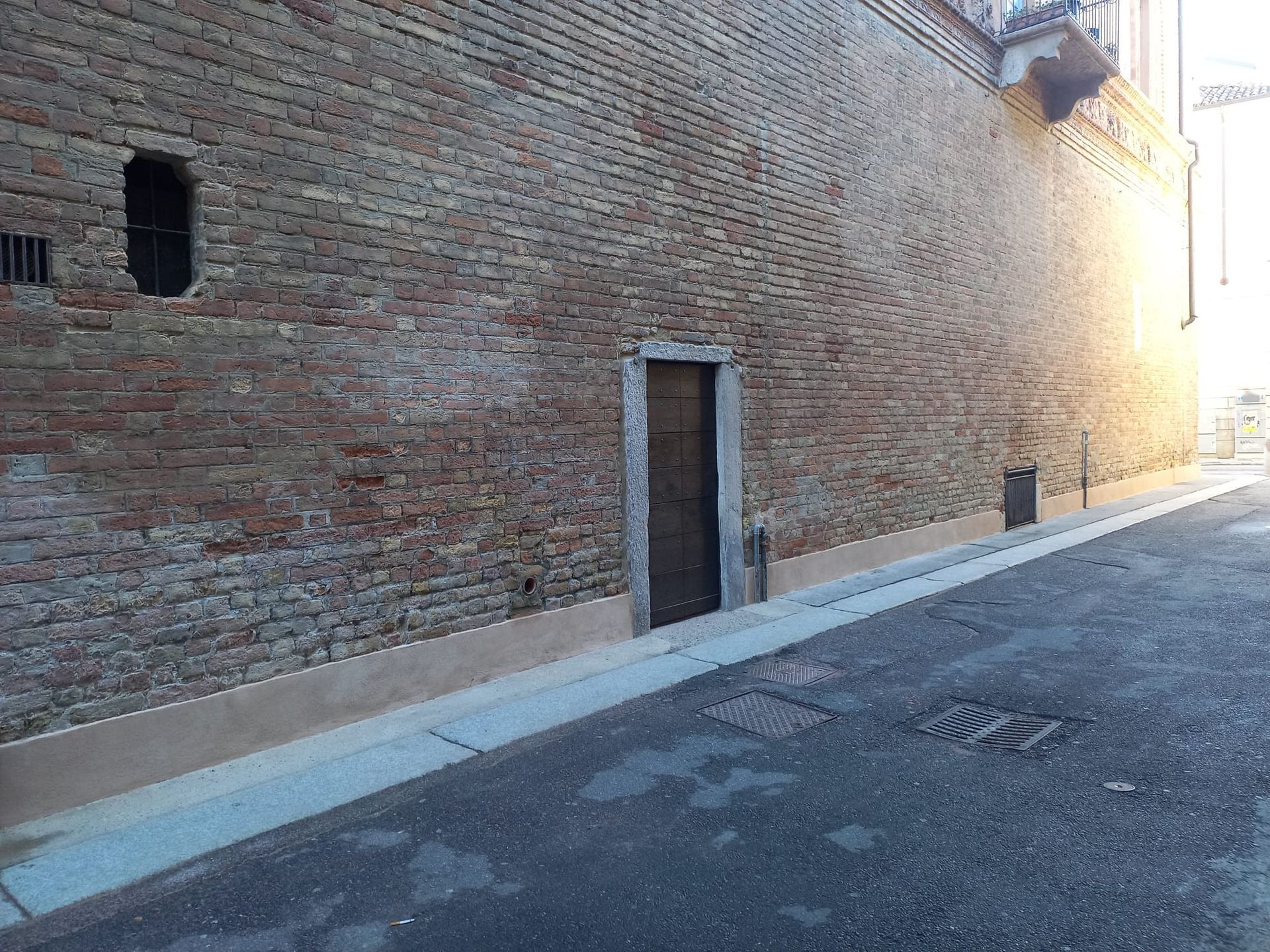 Muro di mattoni con una porta marrone, marciapiede stretto e strada; soleggiato.