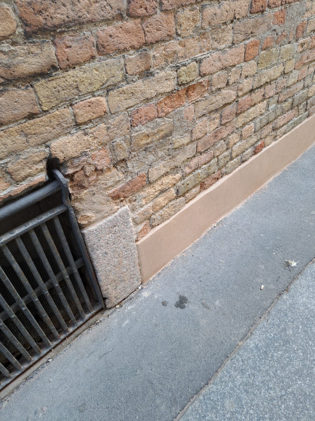 Muro di mattoni accanto a un marciapiede con una grata metallica e un cordolo marrone.