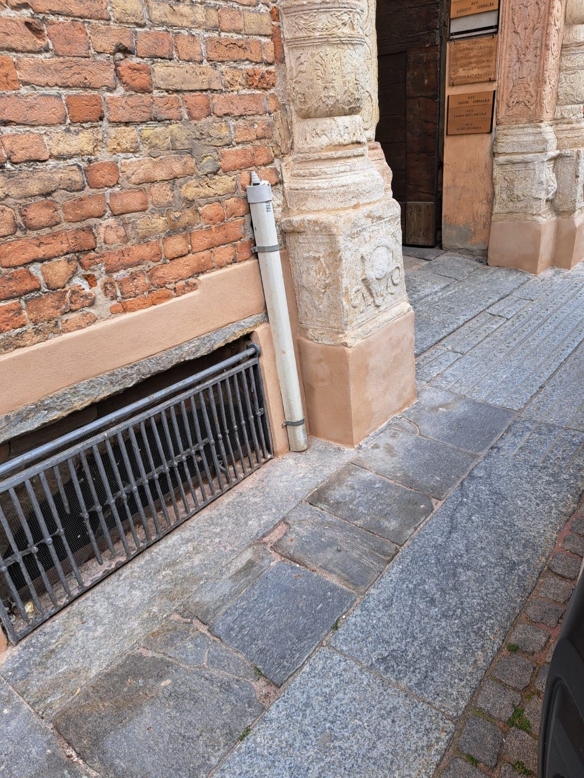 Muro di mattoni con una griglia metallica sul fondo, accanto a un camminamento in pietra. Lungo il percorso corre un tubo di scarico bianco.