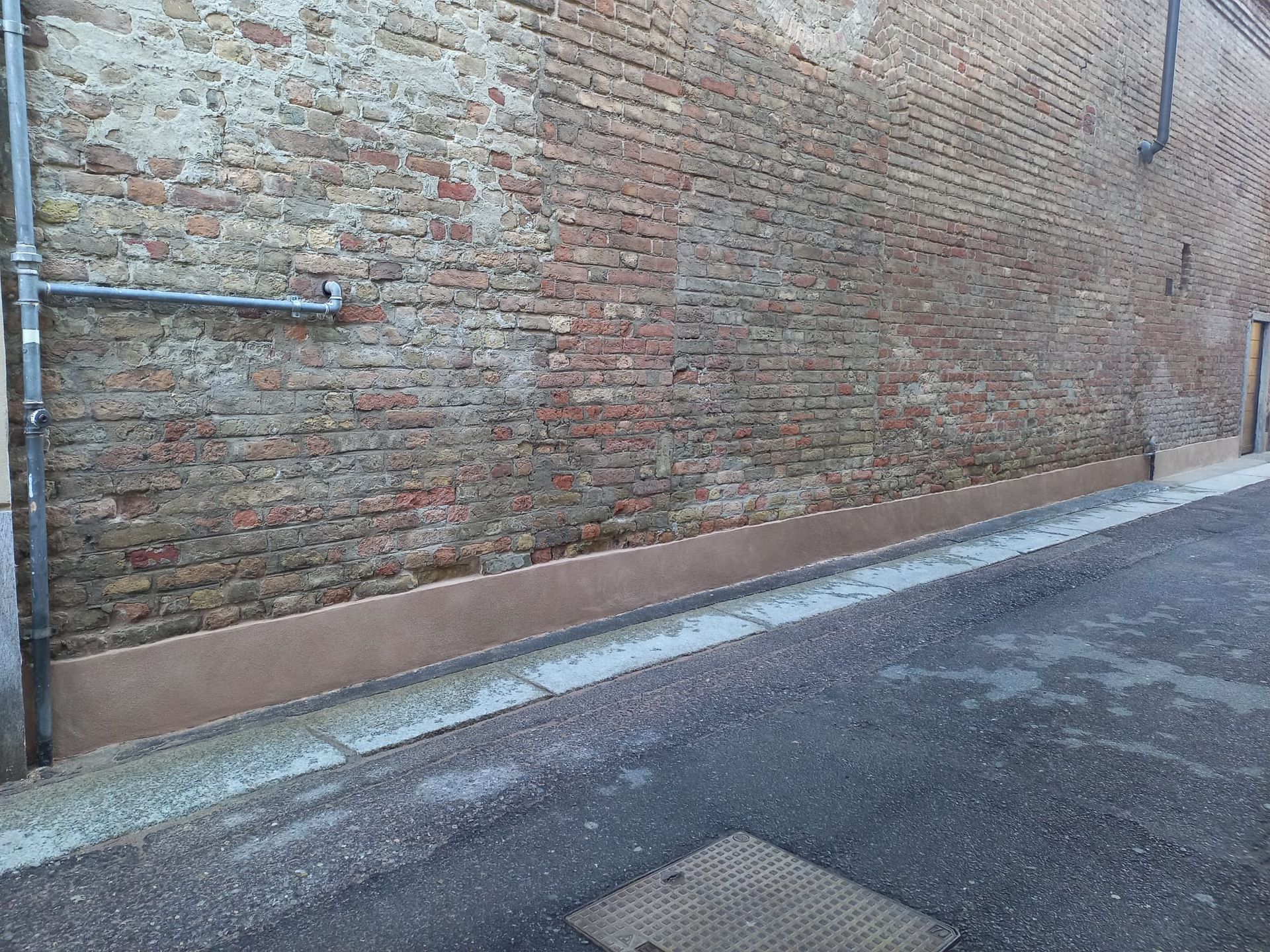 Muro di mattoni con base marrone lungo una strada asfaltata e un tombino metallico.